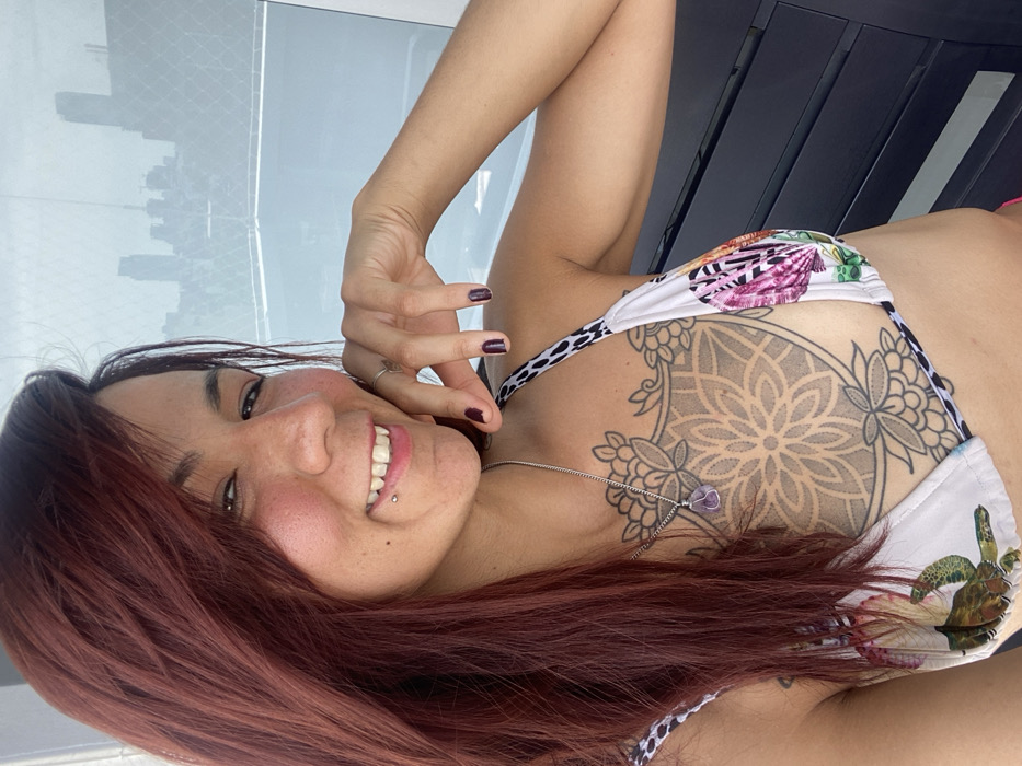 A woman with auburn hair smiles towards the camera. She has a pierced lip and dark nail polish. She wears a bikini top with a floral and shell print. A large, circular tattoo adorns her chest above her breasts, with a necklace resting on top. In the background, a cityscape is visible through a window or opening. Dark furniture partially frames the shot.