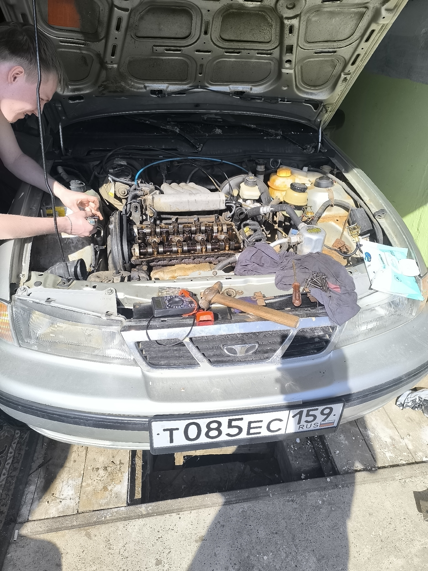 A person is shown working on a car engine in what appears to be a garage. The car is silver, with its hood open, revealing the engine compartment. A hammer, screwdriver, and multimeter rest on the car's front grill. The license plate reads "T085EC 159 RUS", indicating a vehicle registered in the Perm region of Russia. The garage floor has a pit in front of the car.