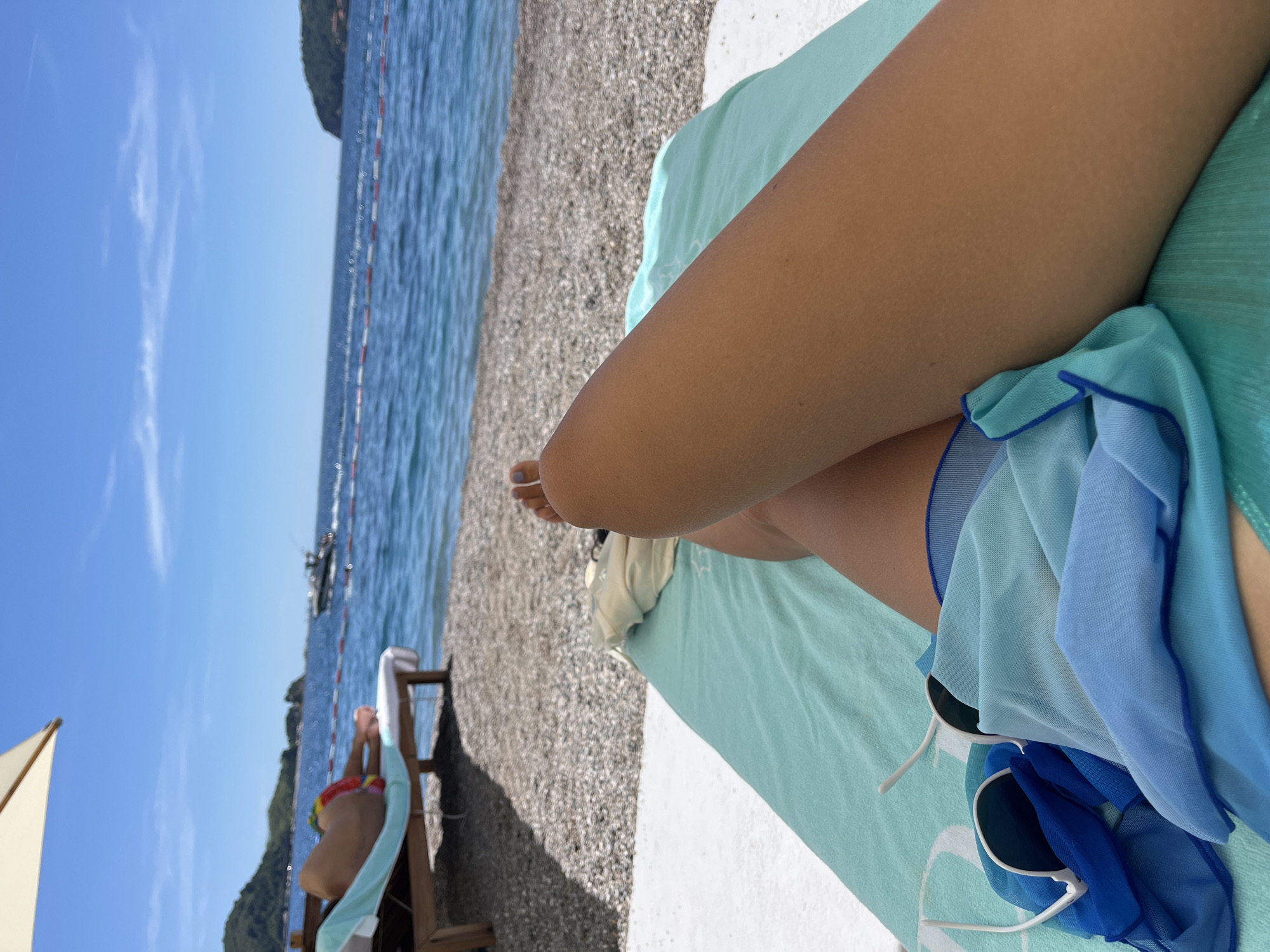 The image captures a first-person perspective of someone relaxing on a beach in Budva, Montenegro. The foreground features a person's legs and a turquoise towel, with a pair of sunglasses and blue fabric draped over it. The background reveals a pebble beach, the clear blue Adriatic Sea, and a distant shoreline with lush green hills and some buildings. A boat is visible on the water, and a beach umbrella provides shade. Another person is seen lying on a sun lounger in the mid-ground. The sky is bright blue with a few wispy clouds.