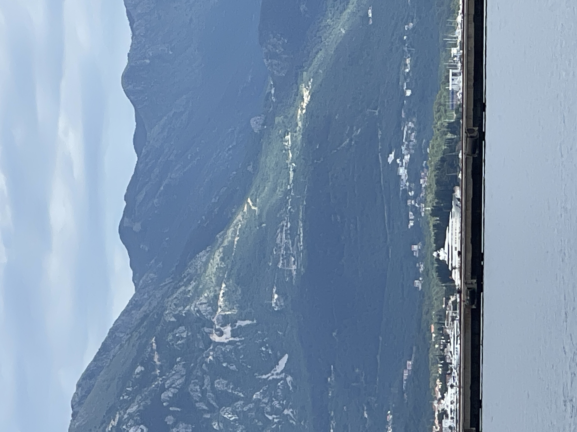 This horizontal landscape photograph captures a scenic view of the mountainous coastline of Bijela, Montenegro. The scene is dominated by a majestic, dark green mountain range that rises steeply from the water's edge, stretching across the middle and left portions of the frame. The mountain slopes are covered in dense vegetation, with scattered lighter patches and streaks, possibly indicating exposed rock formations, sparse growth, or trails carved into the terrain.
Above the mountain, the sky is a clear, light blue with soft, wispy white clouds, suggesting a pleasant daytime setting. In the foreground, on the right side of the image, a calm body of water is visible, likely the Bay of Kotor, extending from the bottom to the top of the frame. A dark, vertical structure, possibly a railing from a boat or a waterfront promenade, runs along the immediate right edge, framing the view.
Along the base of the mountains, nestled between the water and the rising slopes, a coastal settlement is visible. Numerous white and light-colored buildings are scattered amidst green areas, indicating a vibrant town or village. No specific activity or individuals are discernible, presenting a tranquil and expansive landscape. There is no visible text in the image.