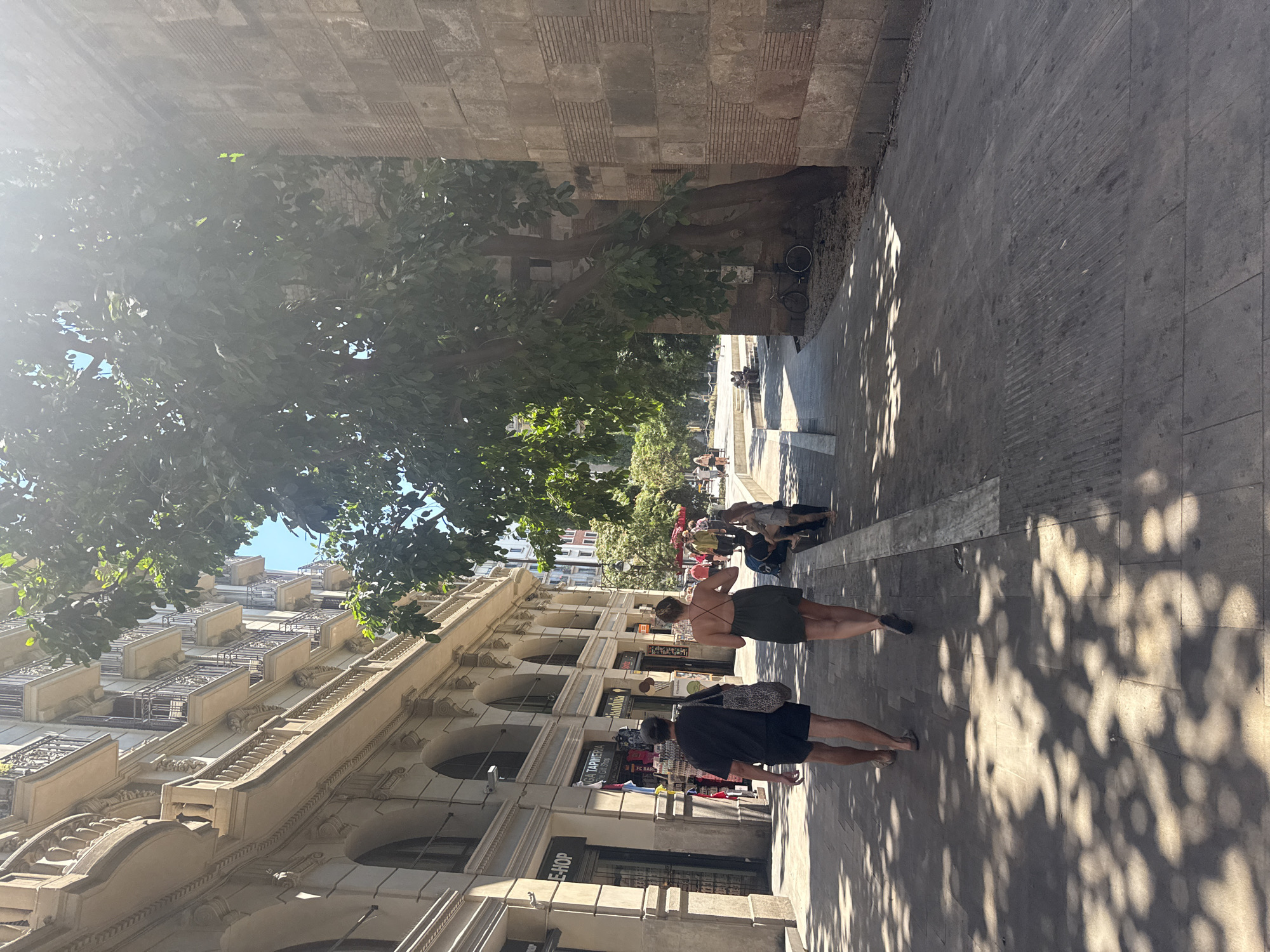 This image depicts a street scene in Barcelona, Spain, likely during the daytime, given the strong shadows cast by sunlight filtering through trees. The perspective is low, looking up towards a building facade and across a sidewalk filled with people.
The foreground shows the legs and torsos of several individuals walking. One person wears a dark green, low-back dress and another a black t-shirt and shorts. They are walking away from the camera on a paved sidewalk. The building on the left is a multi-story structure with ornate architectural details, including arched windows and balconies. Sunlight creates dappled patterns on the sidewalk.
Further down the street, more people are visible, some appearing to be seated or standing in groups. Trees with lush green foliage are prominent, casting shade. A bicycle is parked near the base of a large tree and an old stone wall. The background reveals more buildings and a bright blue sky.
Visible text includes "SHOP" on a sign on the building facade, and "E-HOP" and "FC BAR" are partially visible on other signs. The overall atmosphere appears casual and lively, suggesting a public space in a city. The orientation of the image is rotated 90 degrees clockwise.