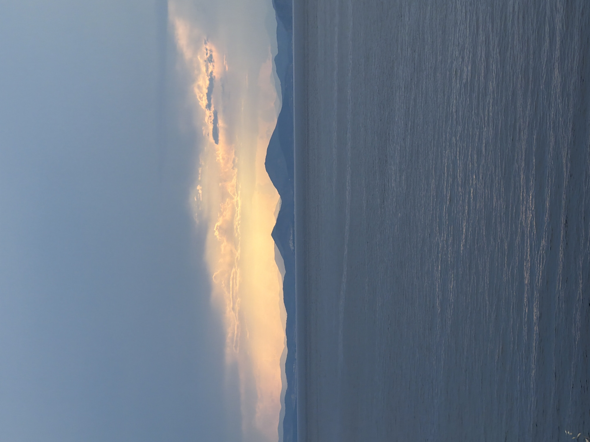 The image shows a serene landscape, seemingly taken near Hydra, Greece, given the presence of a body of water and mountainous terrain. The scene is vertically oriented. The right portion of the image is dominated by calm water, its surface showing subtle reflections of light. Along the horizon, a series of mountains stretch across the frame, their silhouettes fading into the distance. To the left of the mountains, the sky is filled with clouds, partially illuminated by what appears to be the setting sun, casting a soft, golden glow. The overall impression is that of a quiet, natural setting captured at dusk or dawn.