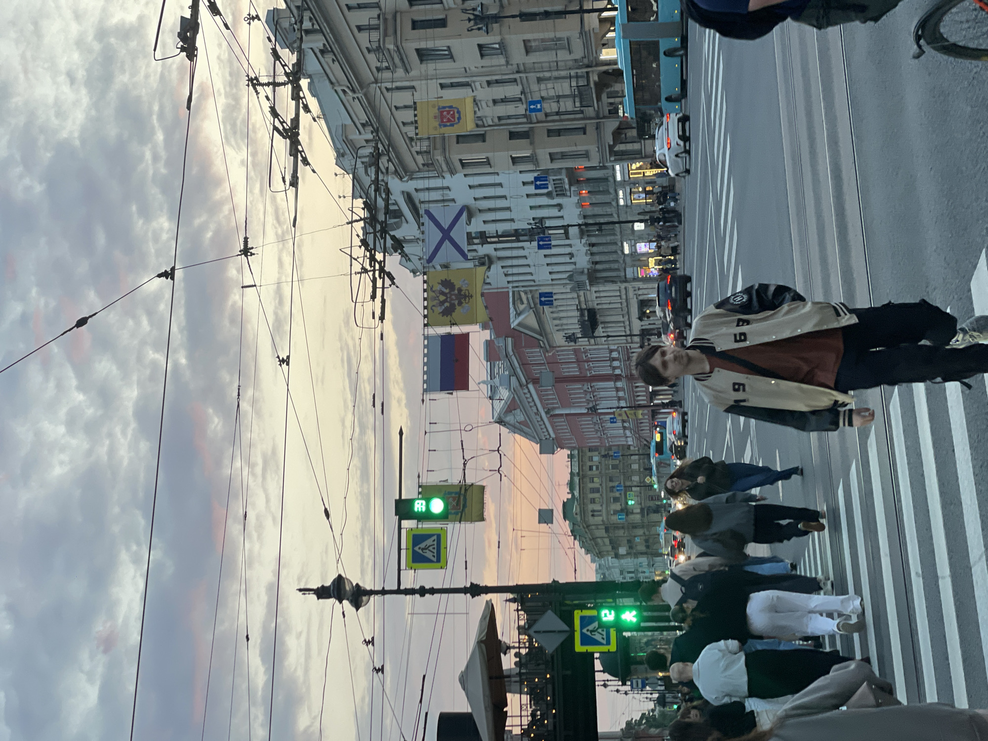The image depicts a street scene in Saint Petersburg, Russia, featuring a pedestrian crossing. Pedestrians are crossing the street, including a person in a beige and black jacket with the number "69" on it. Buildings line the street, adorned with flags including the Russian tricolor and other historic banners. Traffic signals display a green light for pedestrians. Power lines and light posts are also visible. The sky suggests either sunset or sunrise.