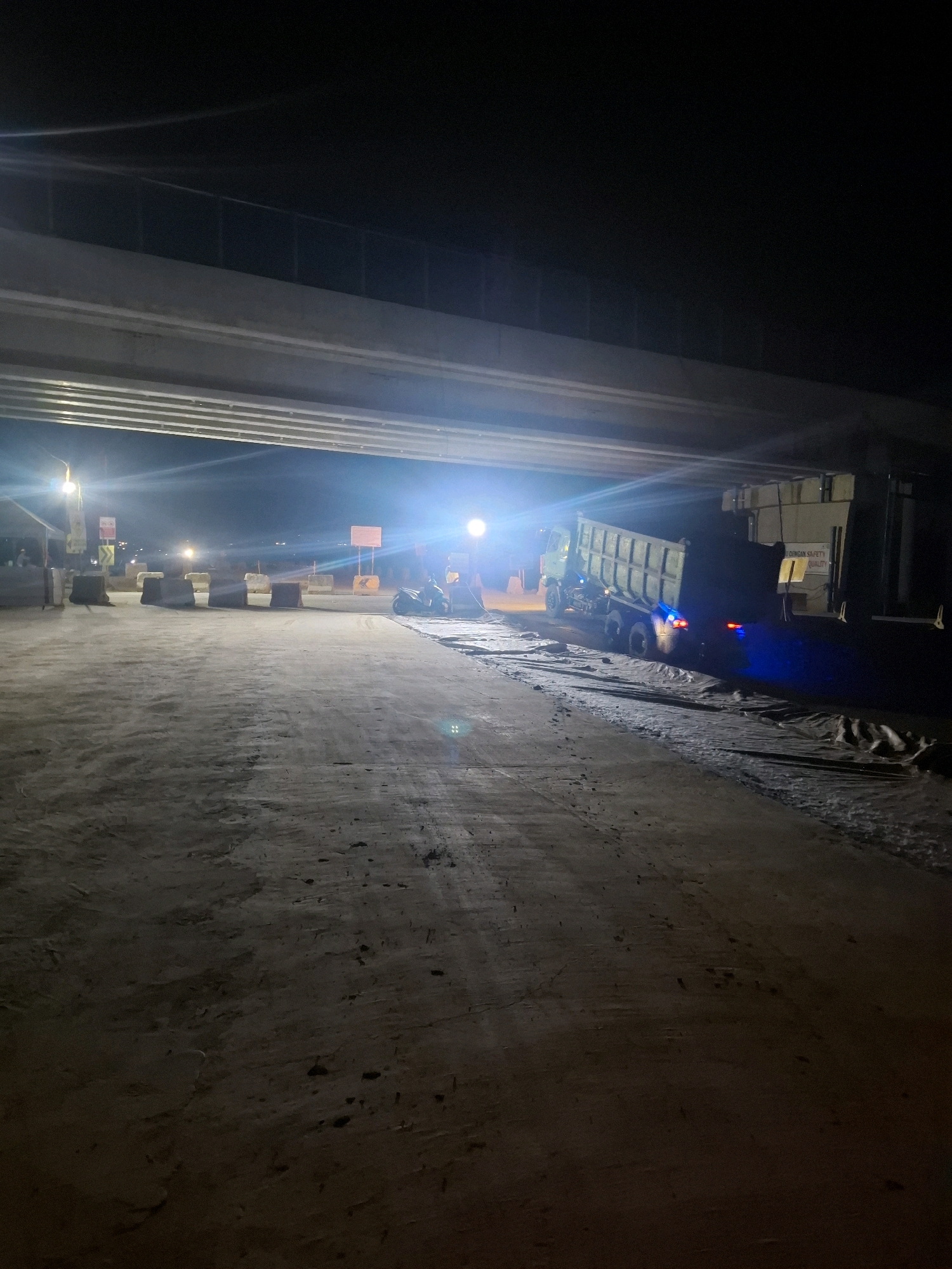 The image depicts a construction site at night, under an elevated structure. The ground is unpaved and appears to be dirt and gravel. In the mid-ground, a large dump truck with its bed raised is visible. The truck is illuminated by bright lights, casting blue and white light onto the surrounding area. Several concrete barriers are positioned across the site, likely for traffic control or to delineate work zones. In the background, there are additional lights, signs, and what appears to be a small temporary structure or booth. The overall atmosphere is one of ongoing construction work at night. The image is taken in Kutasirna, Indonesia.