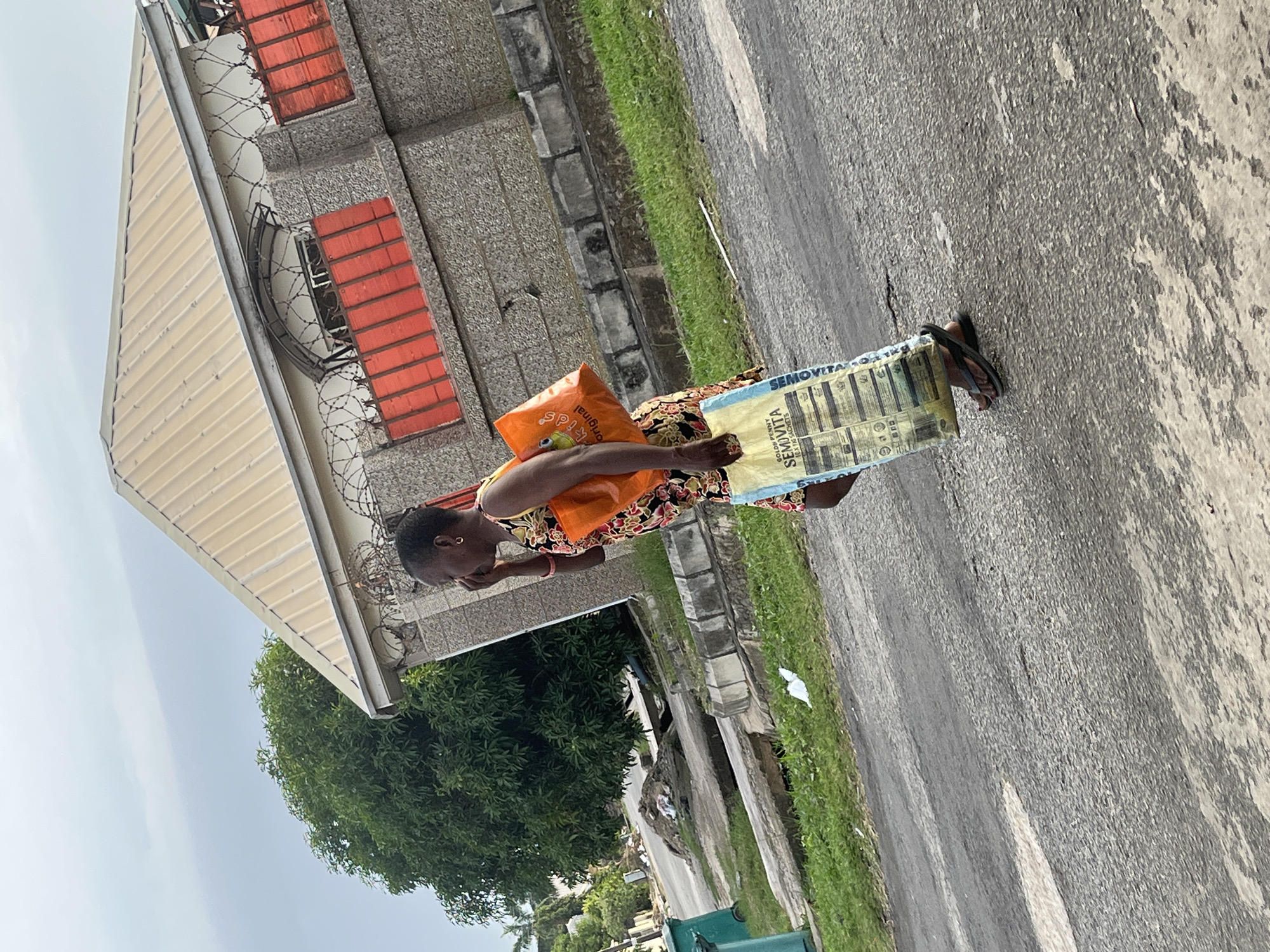 A woman is captured from the side, carrying two bags of groceries as she walks on an asphalt road in what appears to be a residential area in Abuja, Nigeria. She is wearing a colorful floral-patterned dress and black flip-flops. One bag, an orange plastic bag, has "Sunlight Kids" printed on it. The other bag is a large, light yellow woven bag with "SEMORITA" printed in blue on it, along with text that appears to be in English, possibly listing ingredients or product details.
The scene includes a building with a light-colored corrugated metal roof and grey textured walls, accented with orange window frames. Barbed wire is visible along the top of the building's wall. To the left of the building, lush green trees are abundant, suggesting a tropical or subtropical climate. The road is bordered by a strip of green grass and a concrete curb. The sky is overcast, indicating a cloudy day. The overall impression is a common, everyday scene of someone returning home with groceries.
