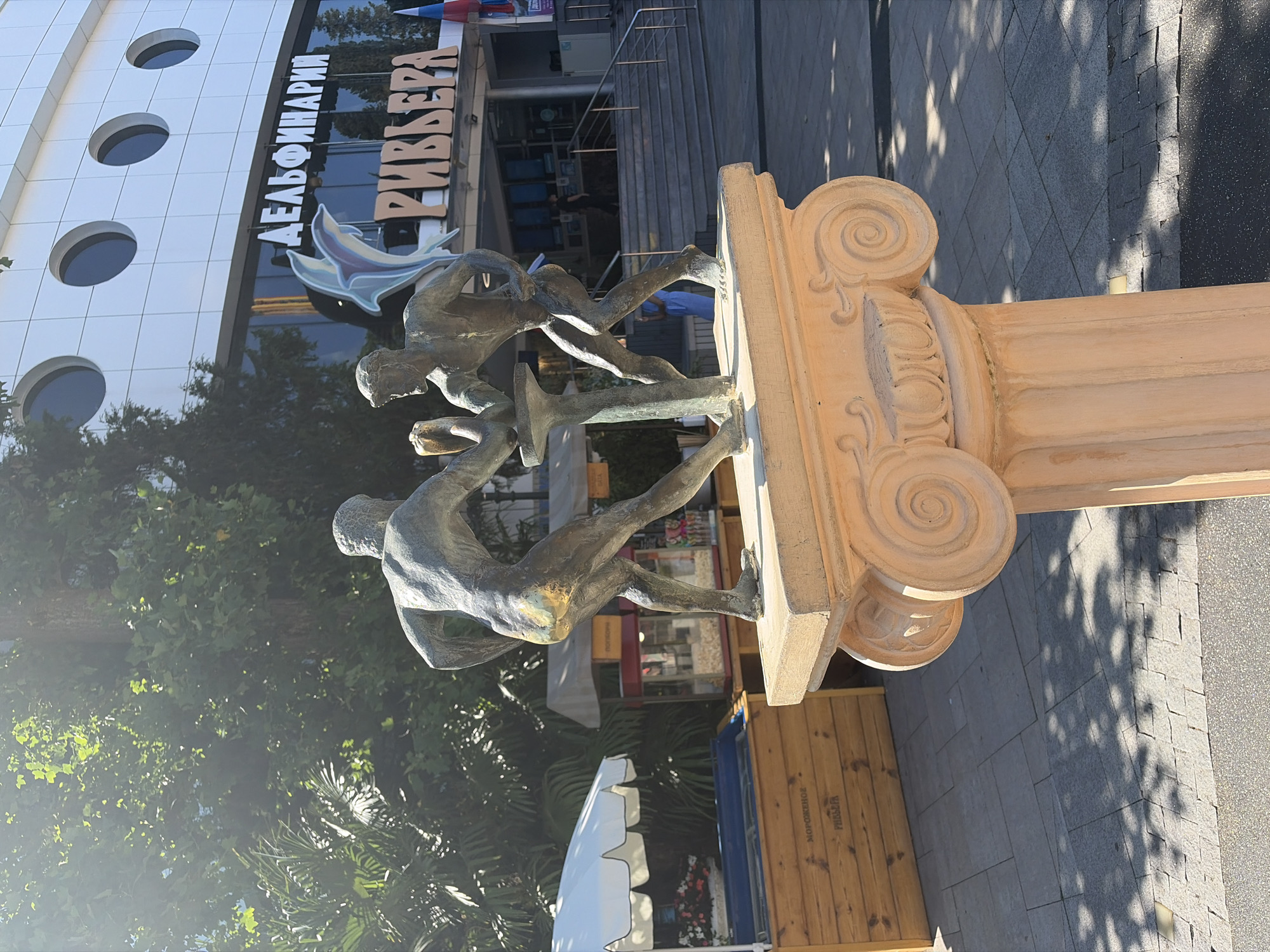 The image depicts an outdoor scene in Sochi, Russia, specifically in front of the Dolphinarium Rivyera. In the foreground, a bronze sculpture of two muscular, nude men engaged in an arm-wrestling match stands on a pedestal adorned with Ionic volutes. The pedestal is light orange and weathered. Behind the sculpture, the Dolphinarium Rivyera building is visible, characterized by its white facade with circular windows and the prominent Cyrillic signage "ДЕЛЬФИНАРИЯ" and "РИВЬЕРА". A stylized dolphin emblem is also visible on the building.
To the left of the sculpture, there are wooden kiosks, one with a white umbrella canopy and flowers, and another with a popcorn machine. The ground is paved with light-colored square tiles, with shadows of trees cast upon it, suggesting a sunny day. In the background, lush green trees and palm fronds are present, with a few people visible on the steps leading into the Dolphinarium. The overall impression is of a public recreational area with a focus on entertainment and attractions.