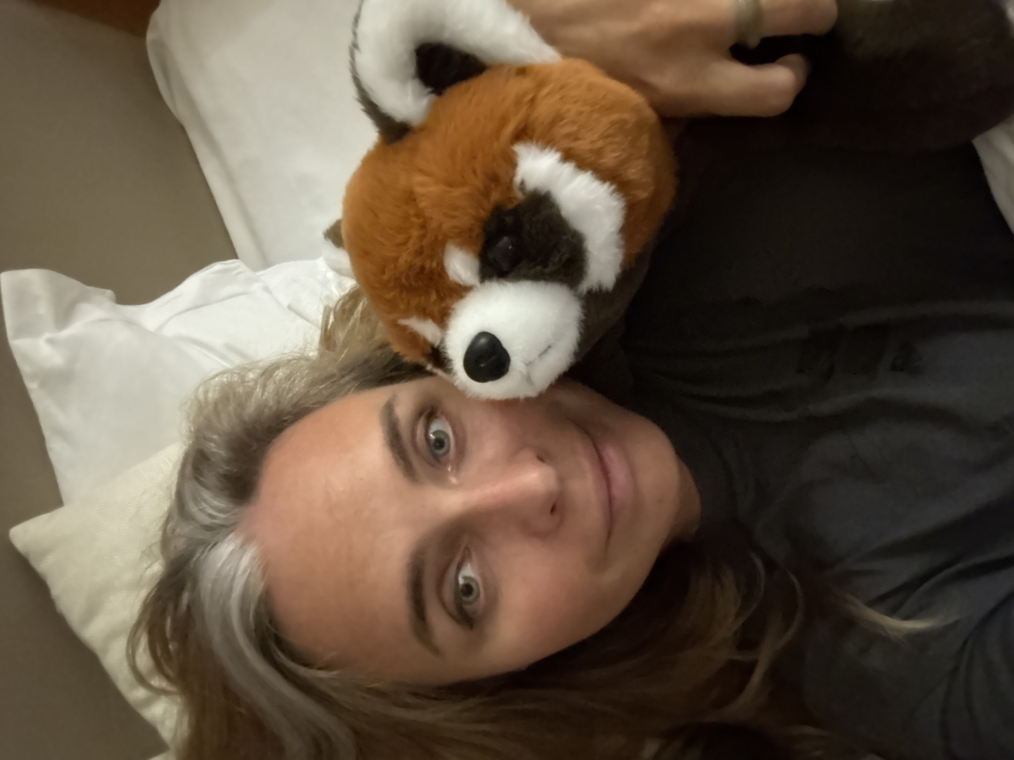 The image shows a close-up, selfie-style shot of a woman with graying brown hair lying in bed. She is holding a stuffed red panda plush toy to her cheek. The woman has green eyes and is wearing a dark t-shirt. Behind her, white pillows and bedding are visible. The lighting is soft and appears to be indoor. The location is not specified in the image or the prompt, and therefore cannot be determined.