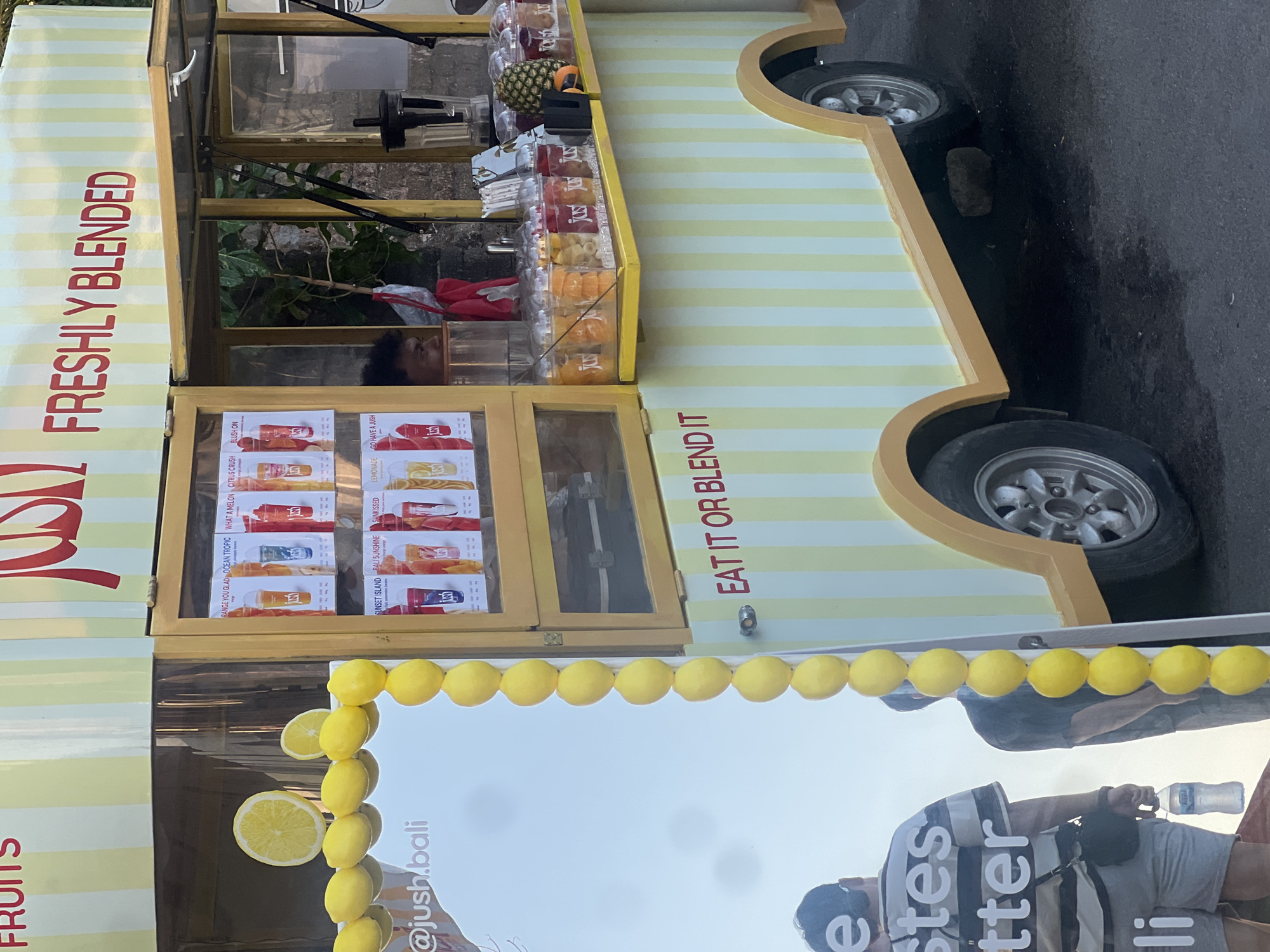 An outdoor juice cart, likely in North Kuta, Indonesia, is adorned with yellow and white stripes. The cart displays various juices in clear cups, labeled with different fruit names such as "Ocean Tropic" and "Citrus Crush." Lemons are a prominent decorative element, with a large slice of lemon and several whole lemons displayed on the front of the cart and a string of lemons cascading down the side. A man can be seen behind the counter, interacting with customers or preparing drinks. In the foreground, a person in a striped shirt and shorts holds a water bottle, suggesting a casual, public setting. The cart's signage includes the brand name "JUSH" and the slogan "FRESHLY BLENDED." The ground is asphalt, with some wet patches, indicating it may have rained recently or the area is prone to moisture.