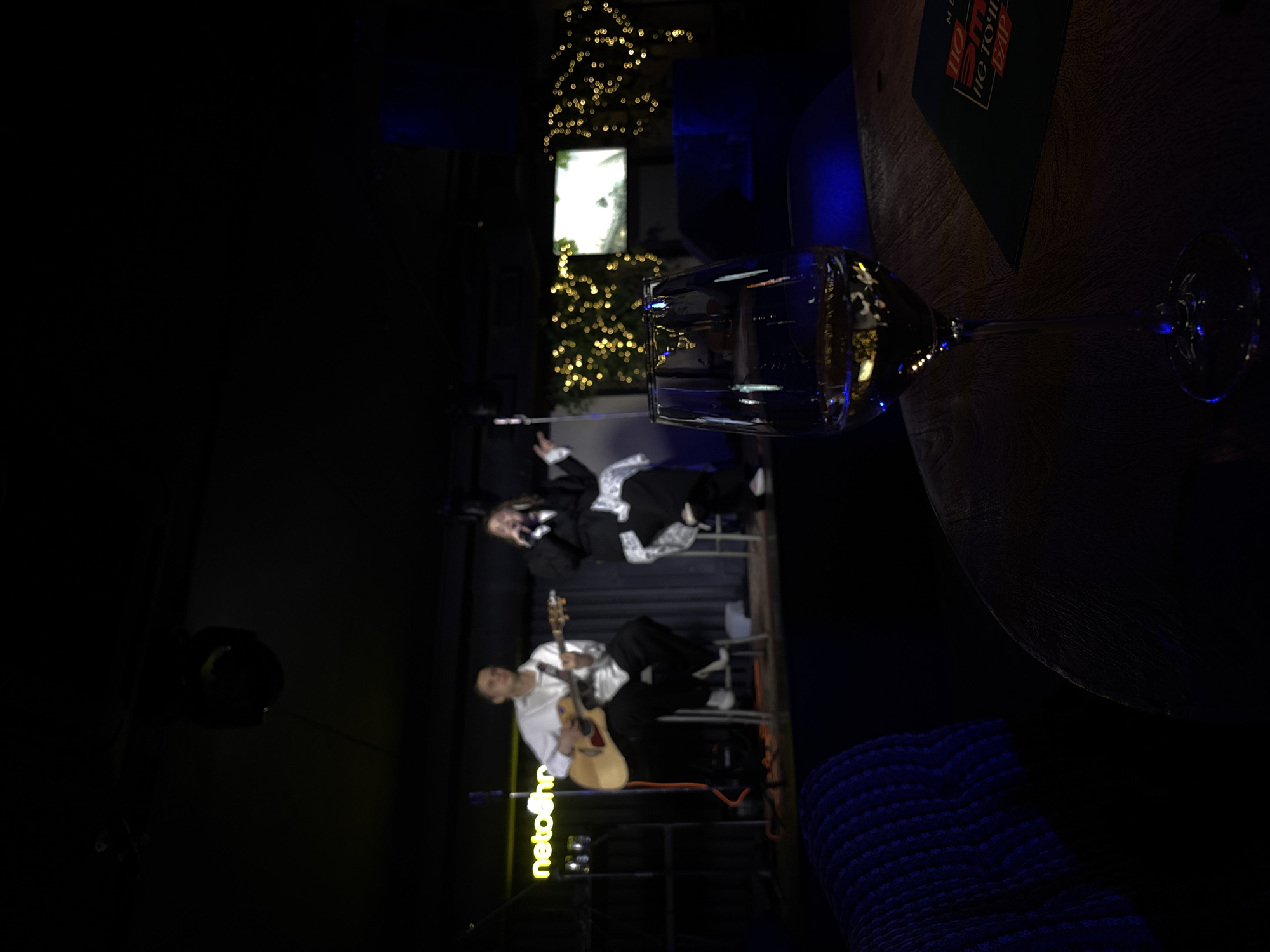 A slightly rotated, low-light indoor shot features a wine glass in the foreground. Behind the glass, there is a stage with two performers: a seated guitarist in a white shirt and a standing vocalist in a black suit with white trim. Behind them is a backdrop of Christmas lights and a window. A sign with the word "netolhr" in yellow neon is visible near the guitarist. In the upper right corner, a menu is visible. The location is likely a bar or restaurant in Krasnoyarsk, Russia. The overall atmosphere suggests a live music performance in an intimate setting.