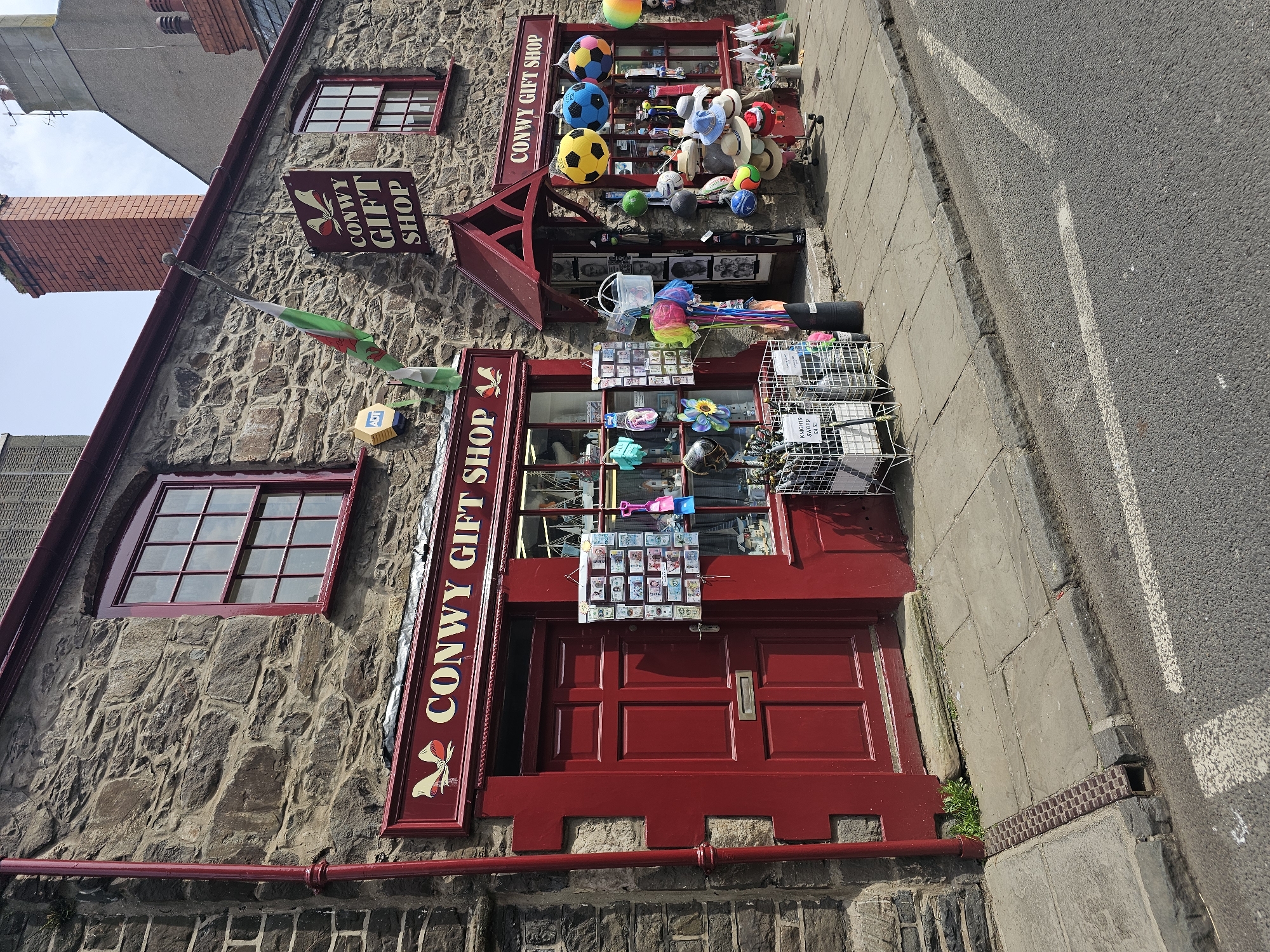 The image depicts the facade of the "Conwy Gift Shop," a two-story building with stone walls and red trim, located in Llandudno Junction, United Kingdom. The shop's signage is prominently displayed on both floors. Souvenirs, including balls, hats, and postcards, are exhibited in front of the shop windows and entrance. An ADT security sign is visible on the wall. A Welsh flag hangs near the upper window. The adjacent pavement and road are visible, indicating an urban setting.