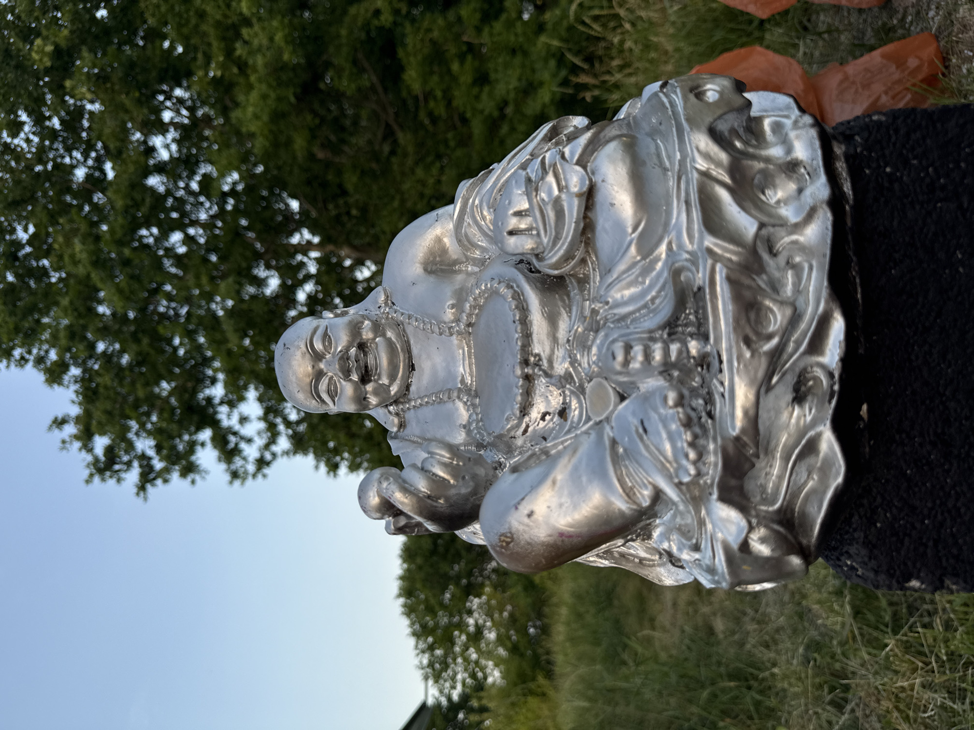 A silver statue of a laughing Buddha sits outdoors in Vol'noe, Russia. The statue is on a dark, textured pedestal. The Buddha is depicted in a seated position with one hand holding a round object and the other resting on its lap. It wears a beaded necklace and ornate robes. The background consists of green grass, trees, and a clear blue sky.