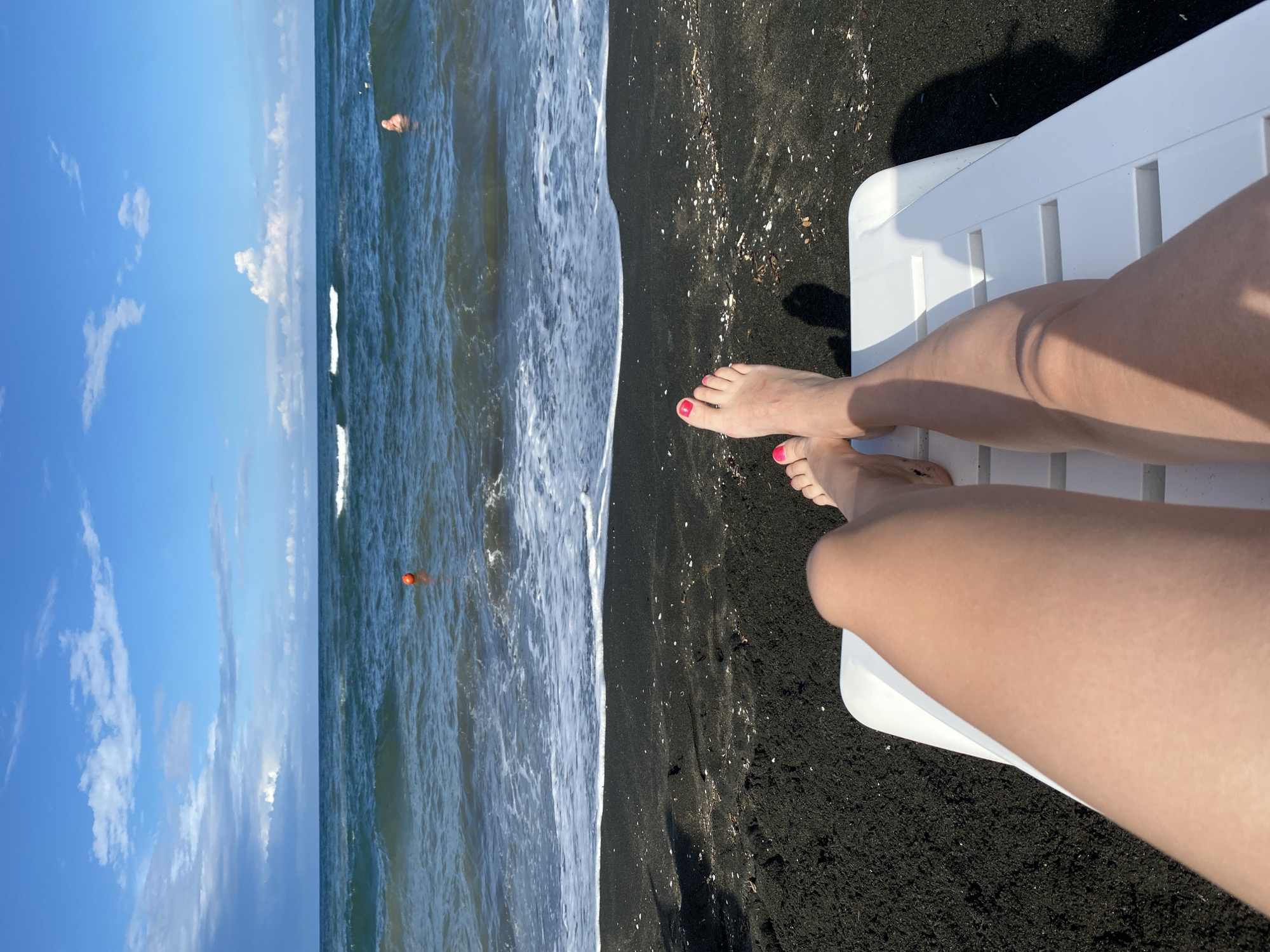 In Ureki, Georgia, a person is relaxing on a white lounge chair on a black sand beach, with their legs extended towards the camera. The person's feet, painted with bright pink nail polish, are visible at the top of the frame. Beyond the beach, the dark blue sea stretches out, with gentle waves lapping at the shore. Two orange buoys are visible in the water, and a person can be seen swimming in the distance. The sky above is a bright blue with scattered white clouds. The overall scene depicts a peaceful day at the beach.