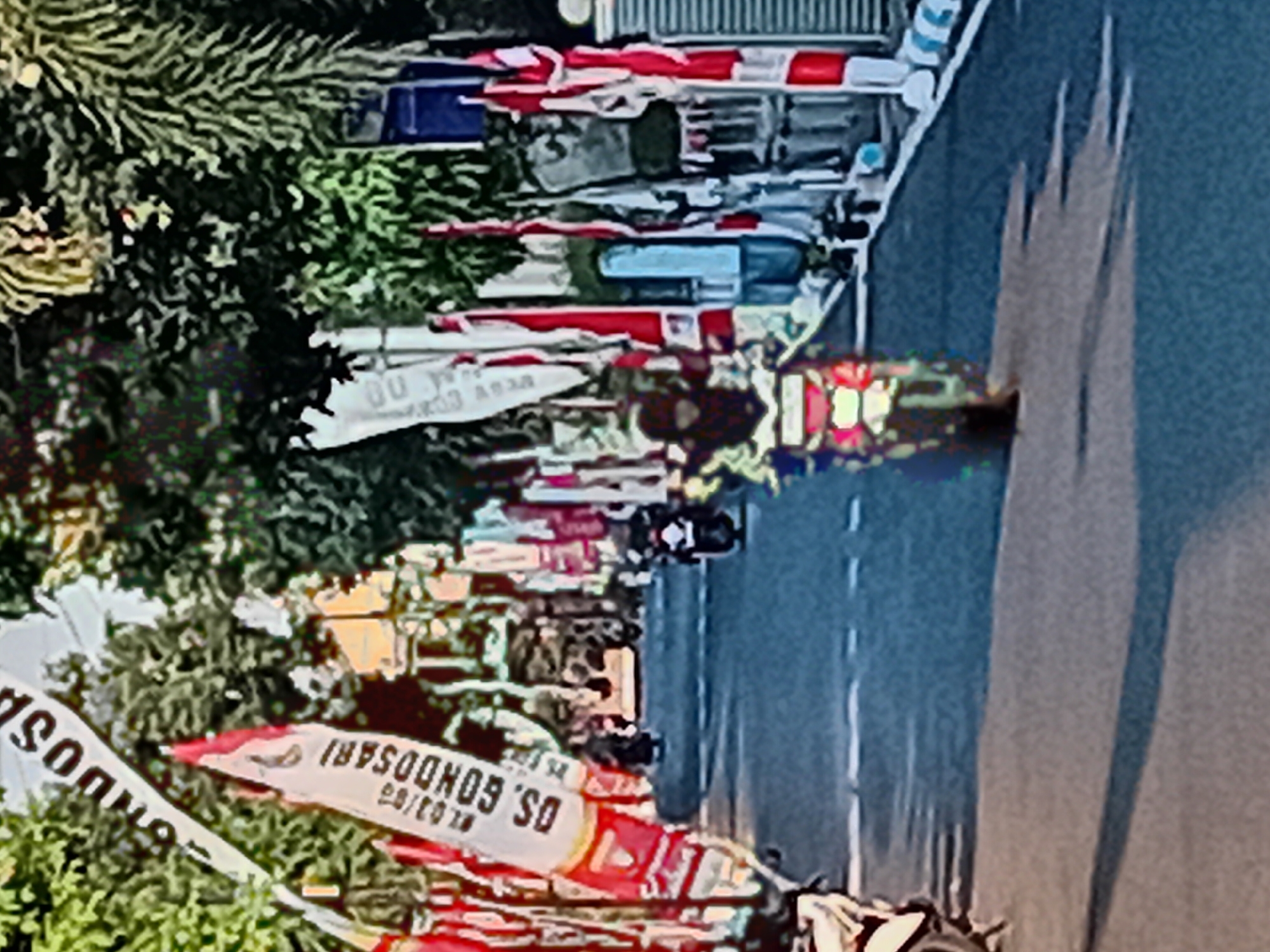 The image captures a street scene in Gebog, Indonesia, decorated with red and white flags and banners. In the foreground, a person on a motorcycle is riding down the road. The motorcycle has a bright headlight, and the rider is wearing a helmet and appears to be a woman. Several other motorcycles are visible in the background, also in motion. The street is paved and has a dashed white line marking the lanes. Trees and buildings line the sides of the street, with some of the buildings appearing to be commercial establishments. The flags and banners suggest a festive or celebratory atmosphere, possibly a local event or holiday. The overall lighting indicates it is daytime.
