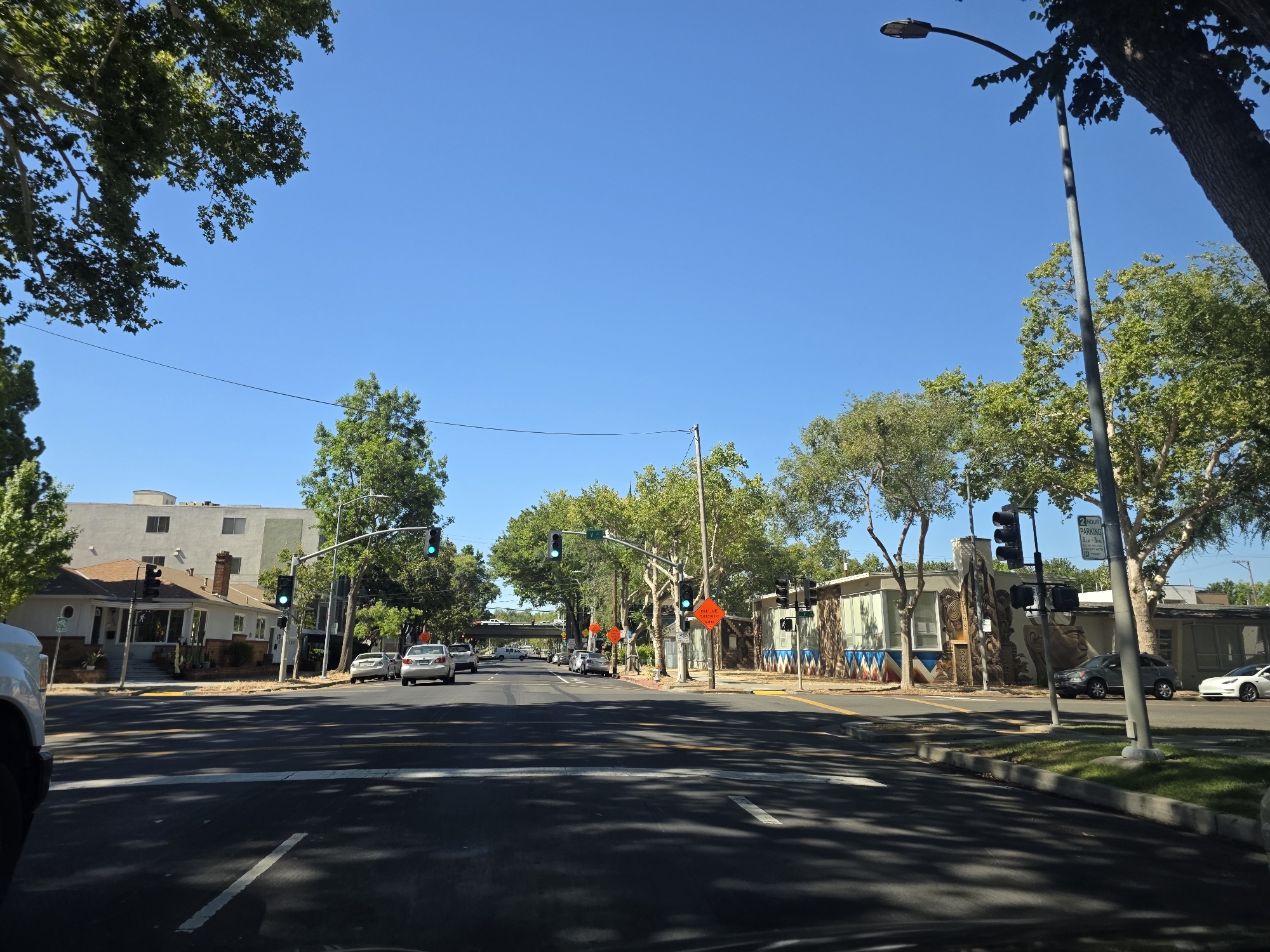 The image depicts a street scene in Sacramento, USA, on a clear, sunny day. The view is from the perspective of a driver, looking down a road lined with trees and buildings. Traffic signals and street lights are visible, some displaying green lights. Several cars are on the road, and some are parked on the side.
On the left side of the street, a single-story house with a pitched roof and a chimney is visible, with a white building in the background. The house has a well-maintained lawn and some potted plants.
On the right side of the street, there are commercial buildings with unique murals and decorative elements. A "2 Hour Parking" sign with specific time restrictions is mounted on a pole. The trees are lush and green, providing shade on the road.
The overall atmosphere is one of a typical urban or suburban environment during daylight hours. There are no people clearly visible in the image, and the focus is on the street, vehicles, and surrounding architecture. The presence of traffic signals and cars suggests ongoing, everyday activity. The bright sunlight and clear blue sky indicate pleasant weather.