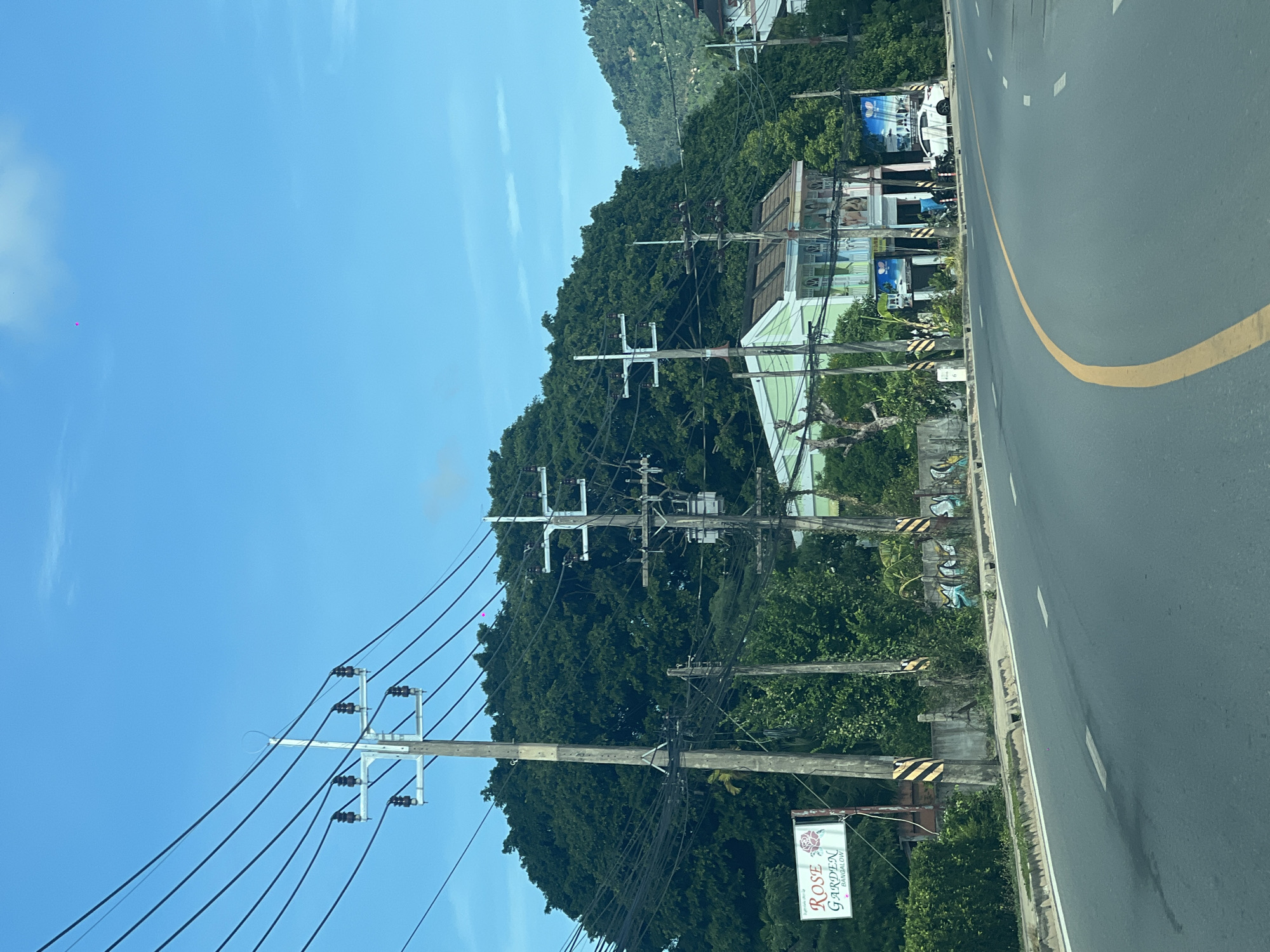 The image depicts a street scene in Baan Chaweng Noi, Thailand, under a clear blue sky. A paved road curves through the frame, marked with white dashed lines and a prominent yellow center line. On the left side of the road, a series of power poles with numerous wires are visible, supporting electrical infrastructure. A sign for "ROSE GARDEN BANGALOW" is mounted on a pole. Behind the road and power poles, a densely vegetated hill covered in lush green trees forms a prominent backdrop. Several buildings are situated along the roadside, including a pale green structure with multiple balconies and windows, and a white car parked in front of one of the buildings. The environment appears to be a semi-urban or suburban area with a mix of infrastructure, natural elements, and commercial establishments.