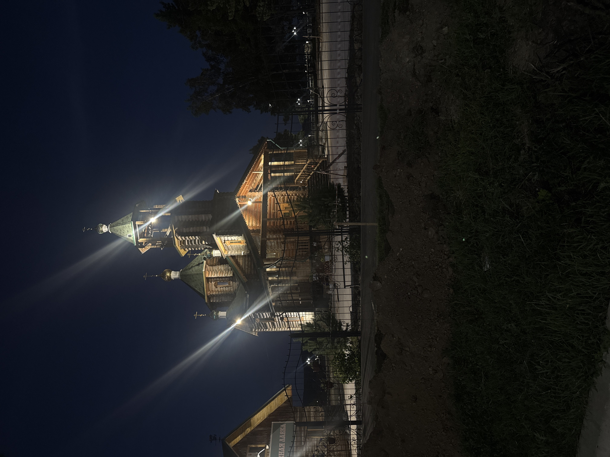 This image captures a wooden church illuminated at night in the city of Komsomolsk-on-Amur, Russia. The architecture features traditional onion domes and cross finials, constructed from logs. The church is situated behind a decorative black metal fence. Adjacent to the church, to the left, is a smaller wooden building with a sign that appears to read "МАГАЗИН" (shop) in Cyrillic script. Streaks of light from strong illumination sources are visible, creating a lens flare effect across the dark sky. The foreground consists of rough ground with piles of dark soil and a strip of grass. In the background, trees are silhouetted against the night sky, and a few small outdoor lights are visible along a pathway behind the fence. The overall scene is one of a quiet, illuminated religious building after dark, with elements of a nearby commercial establishment.