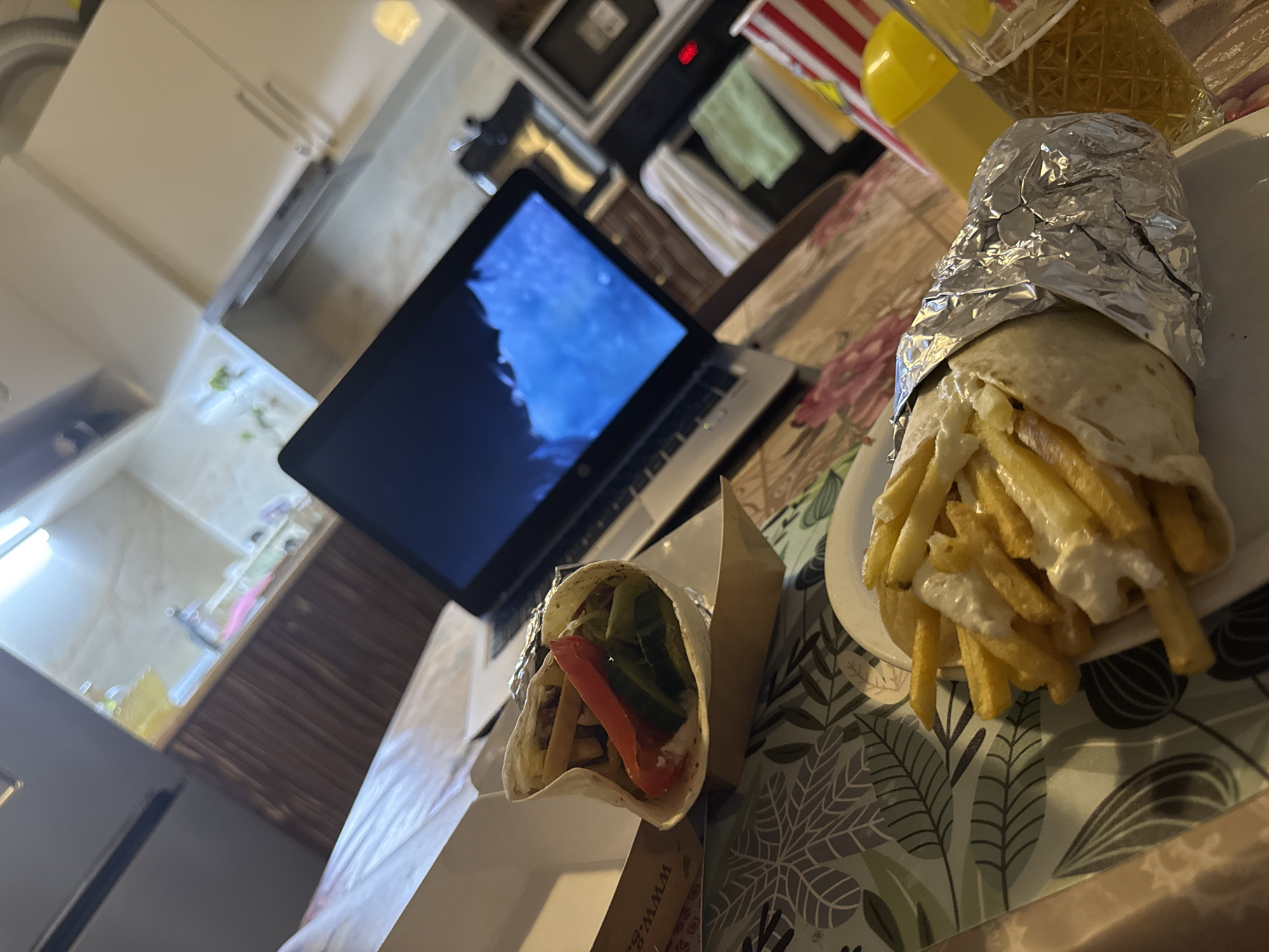 The image shows a close-up view of a meal on a table, possibly in a home kitchen. In the foreground, a wrap filled with french fries and a white sauce is partially covered in aluminum foil. Next to it, on a white plate, is a portion of french fries. A laptop is open in the background, displaying a blue and white image. The table is covered with a patterned tablecloth. A blurred kitchen counter with cabinets and appliances is visible in the background. The overall scene suggests a casual dining experience, potentially while working or relaxing. There is no information in the image to suggest a specific location in Stara Zagora, Bulgaria.