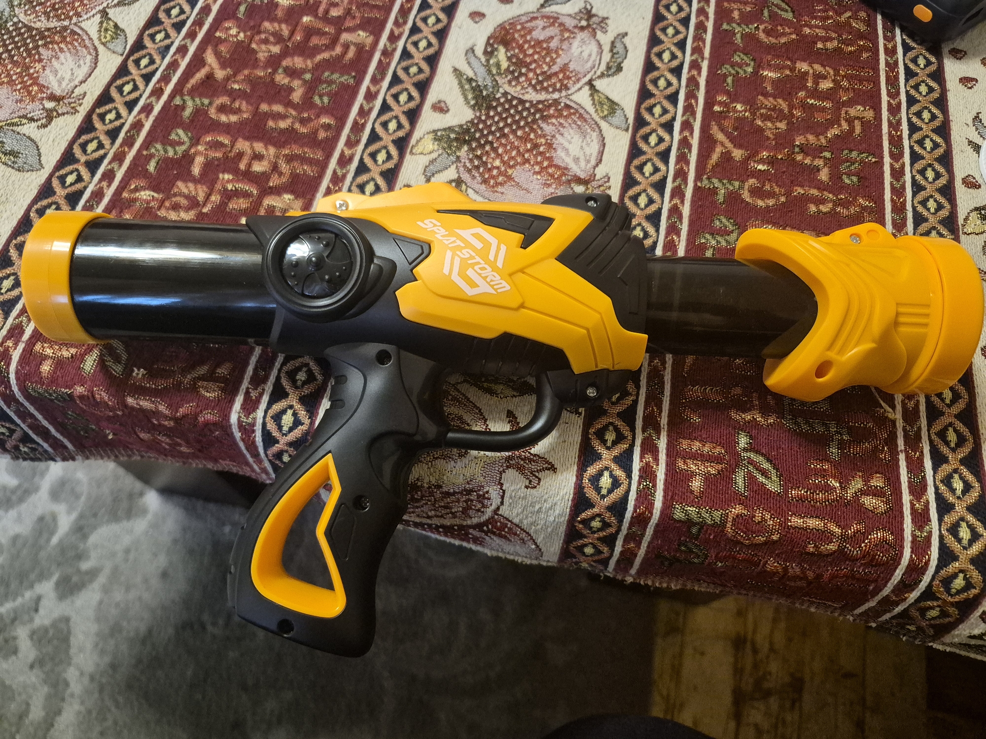 A yellow and black toy bluster, likely a water or gel blaster, is centrally positioned on a patterned textile surface. The bluster features a black body with vibrant yellow accents on the front nozzle, the central mechanism housing, and a portion of the grip and rear. The brand name "SPLAT STORM" is clearly visible in white text on the yellow central section of the toy.
The bluster rests on what appears to be a tablecloth or a decorative fabric, possibly a traditional rug, characterized by a rich, multi-colored design. The dominant color of the fabric is a deep red, interspersed with intricate patterns including cream-colored motifs that resemble pomegranates, and bordered by geometric designs in shades of blue, gold, and white. This style of textile could be indicative of a cultural aesthetic, aligning with the provided context of Yerevan, Armenia.
No people are present in the image, and no specific activity is occurring; the bluster is stationary. The setting appears to be indoors, though the time of day or external weather conditions cannot be determined. Below the patterned textile, a lighter, mottled surface, potentially a floor or carpet, is visible.