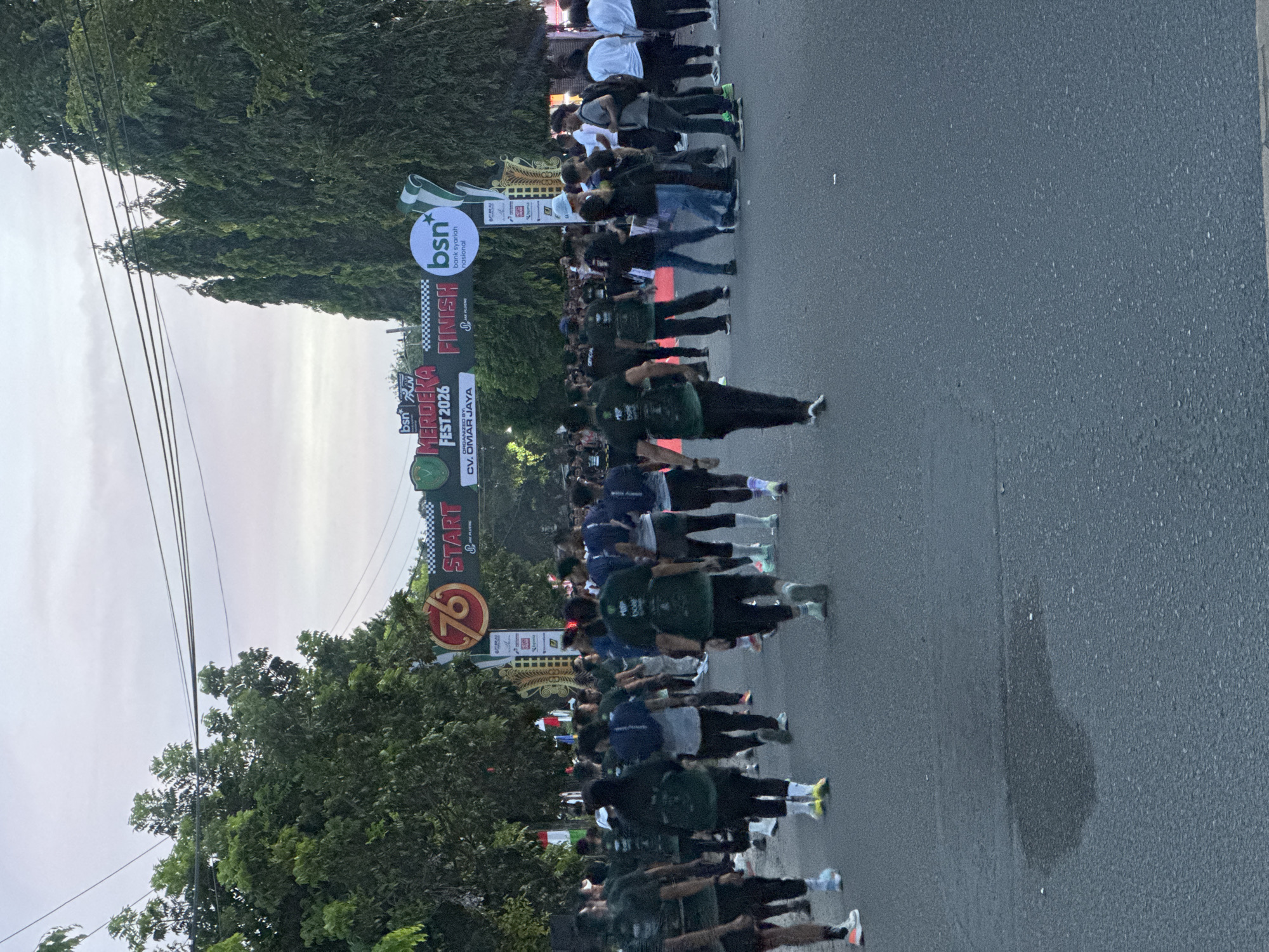 The image depicts a street scene in Lhokseumawe, Indonesia, likely during a running event. A large archway marked "START" on the left and "FINISH" on the right spans the road. The archway is adorned with logos, including "76", "BSN Bank Syariah Nasional", and "MERDEKA FEST 2026". Numerous participants, mostly dressed in dark green shirts, are walking or running on the asphalt road, some approaching the archway and others having passed it. The environment is lined with tall trees and overhead power lines. The sky is overcast, suggesting either early morning or late afternoon. The overall atmosphere appears to be that of an organized public event.