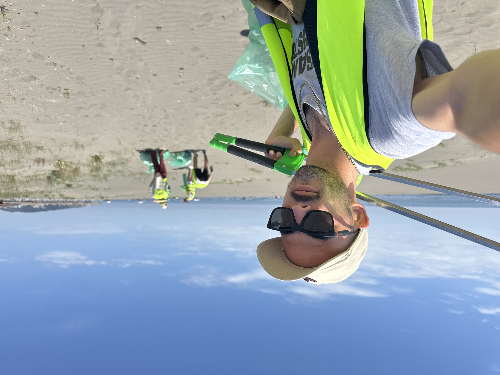 The image shows a man wearing a neon yellow safety vest, a grey shirt, a light-colored cap, and dark sunglasses. He is holding a green and black reaching tool, presumably used for picking up litter. In the background, there are other people also wearing safety vests who appear to be collecting trash. They are on a sandy beach, and there is the sea and horizon line in the distance. The photo seems to be taken as a selfie. The location appears to be the beach near Donji Štoj, Montenegro, based on the geographical features of the coastline and the nature of the beach environment.