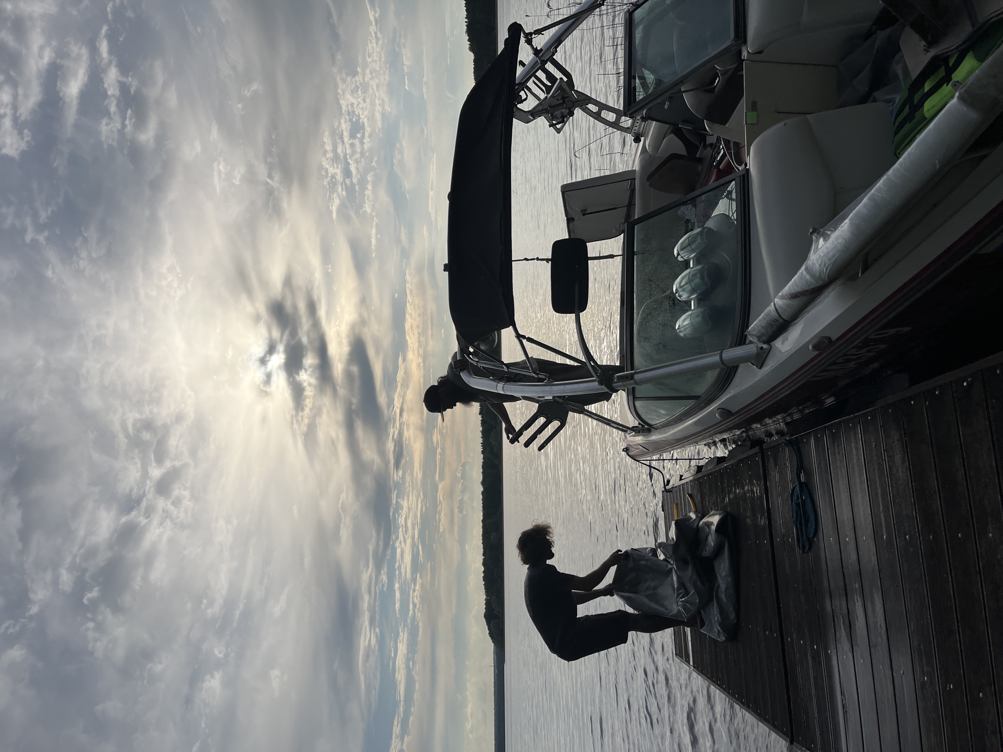 A waterside scene captures two individuals near a boat docked at a pier. One person stands on the dock, holding a large, bundled object wrapped in a reflective material. The other person is on the boat, possibly adjusting equipment or the boat's tower. The boat is moored to the wooden pier, which extends into the water. The sky is filled with clouds, with the sun shining through in the background. In the distance across the water, there is a treeline. Given the landscape and context clues, it is most likely that the image was taken in a rural area in a place such as Пятница, Russia, but there are no identifiable details from the city present in the image.