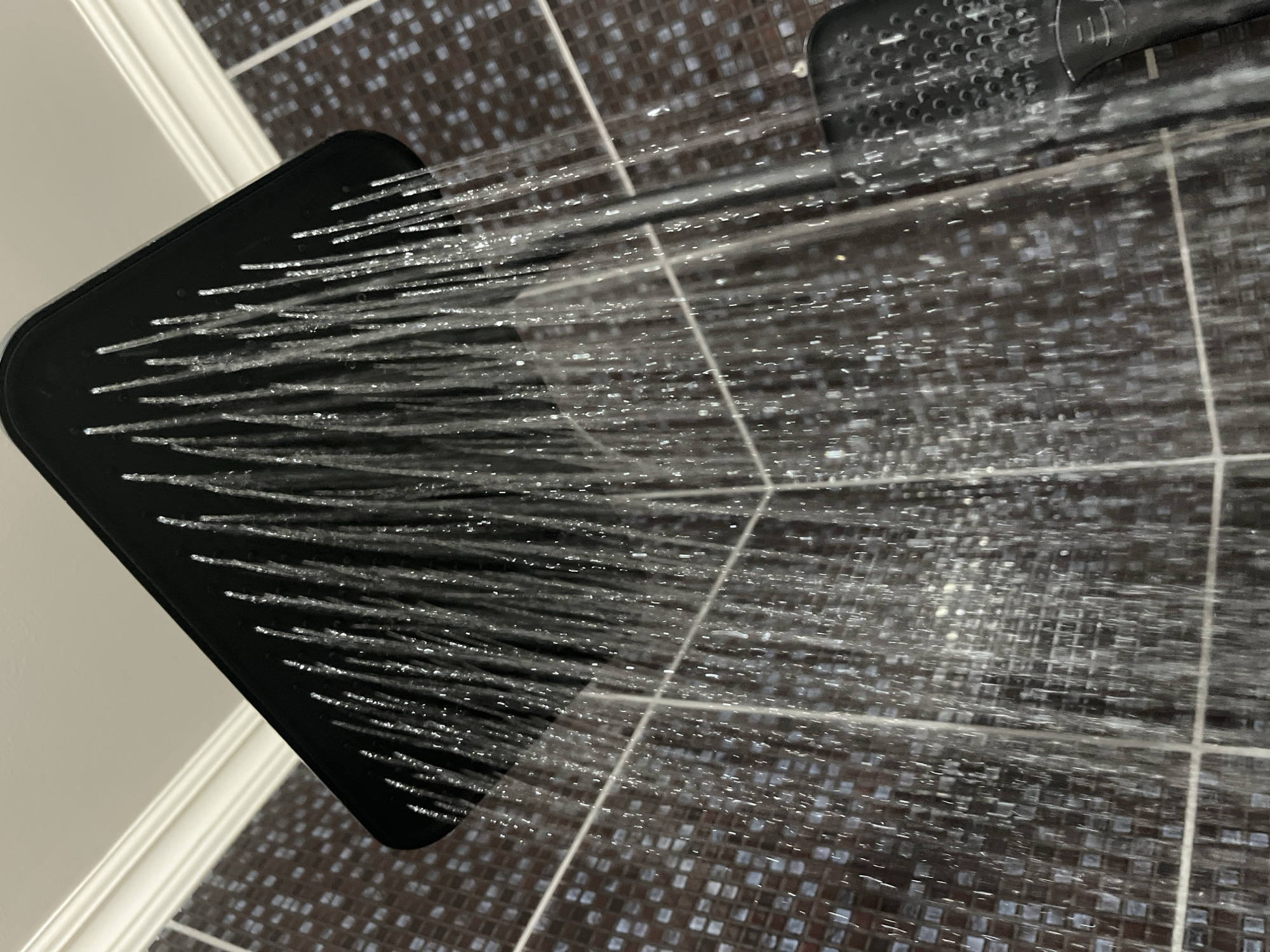 An overhead shot captures a modern black shower head with water streaming down. The shower head is angled downwards, and individual streams of water are visible, catching the light. The background consists of dark, mosaic-tiled walls and a white ceiling with molding. On the right side of the image, part of a hand-held shower head or attachment is visible, also black. The scene is set in a bathroom. No specific location in Shymkent, Kazakhstan can be determined from the image.