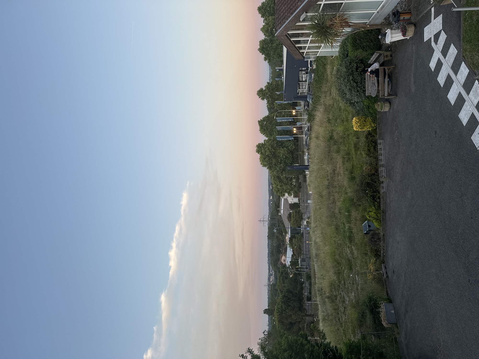 The image depicts an outdoor scene with a clear sky on the left and a subtle gradient transitioning to shades of pink and orange towards the center, possibly indicating sunrise or sunset. Large clouds are present near the transition. On the right, the scene transitions into a suburban environment, featuring a building with large windows and a dark roof, surrounded by greenery. There is a road with marked pedestrian crossings. Closer to the bottom, a mix of trees and foliage is visible. Blue Peugeot dealership flags stand near the building. The image was taken near the Peugeot dealership in Ramonville-Saint-Agne, France.