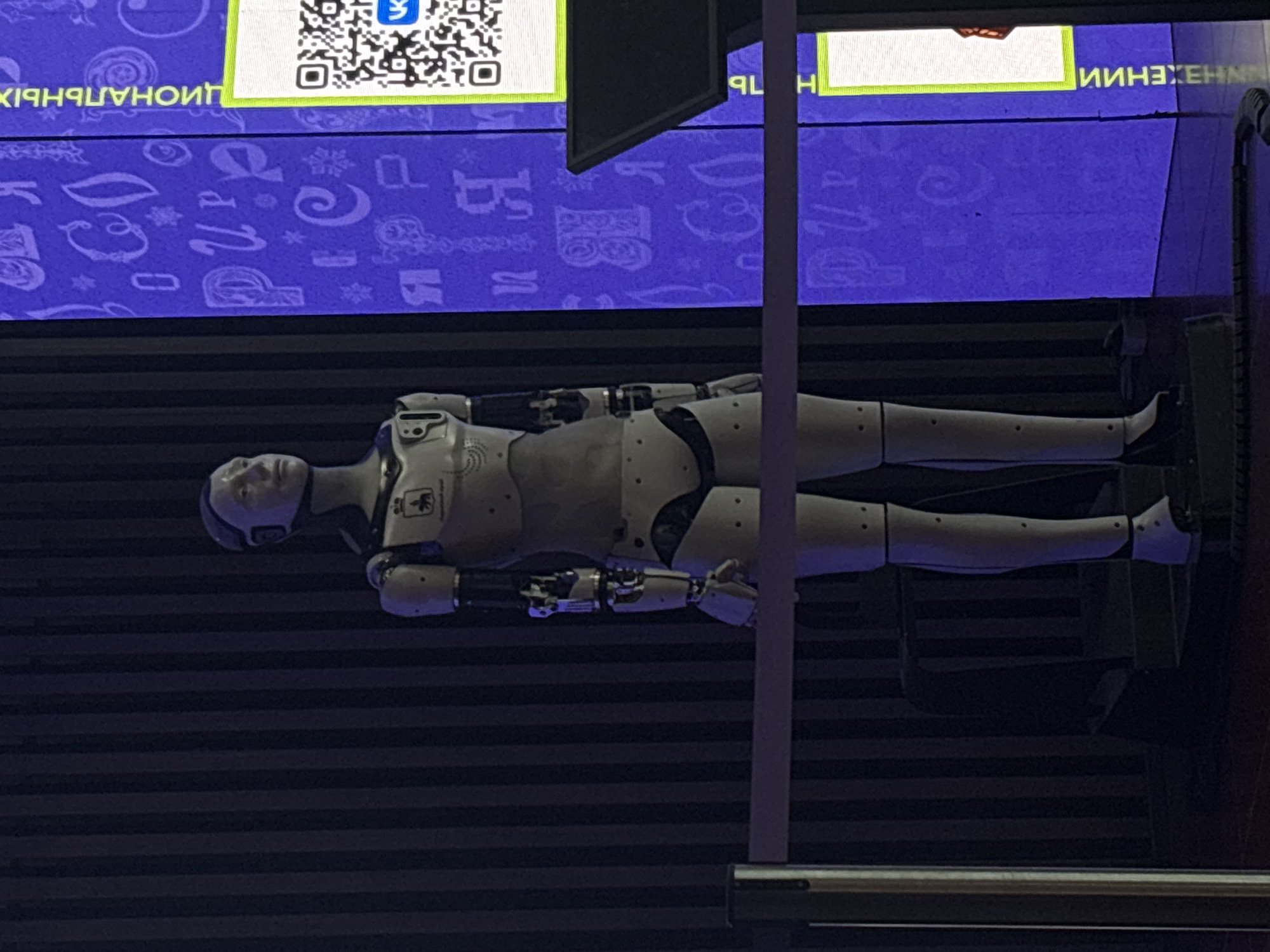 The image shows a humanoid robot lying horizontally against a backdrop that includes a large display screen. The screen features a blue background with white Cyrillic letters and designs. At the top corners of the screen, there are areas with greenish borders and illegible text/symbols within. The robot is white with black accents, and appears to be lying on a dark, striped surface. The robot is in a static pose with its arms positioned near its sides. The location appears to be in Perm, Russia, due to the Cyrillic writing on the screen. A pole is visible vertically in the middle of the image.