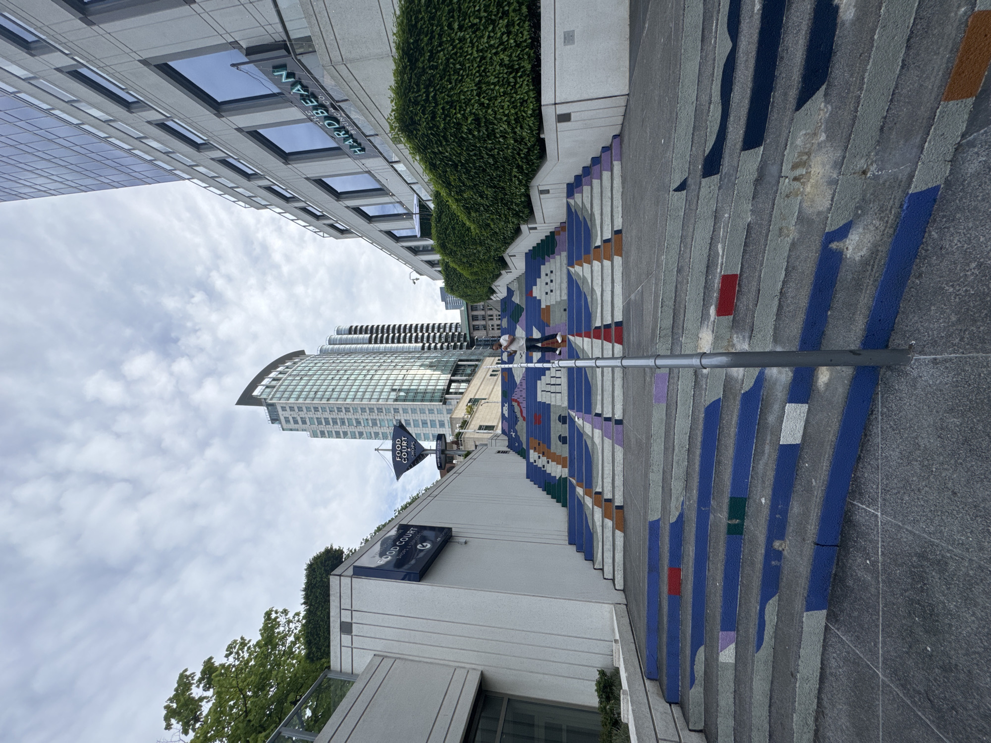 Here is a detailed description of the image:
**Overall Scene:**
The image showcases a set of steps, known as the Vancouver painted stairs, adorned with a colorful geometric design, in Vancouver, Canada. The perspective is a ground-level view looking up the staircase.
**Buildings and Environment:**
To the left of the frame are buildings, including one with the sign "HARD BEAN BRUNCH CO." Another building features a sign for "FOOD COURT." The sky above is partly cloudy. Green foliage and bushes are planted near the buildings and along the staircase.
**Stairs and Art:**
The painted steps are the primary focus, displaying a pattern of blue, grey, purple, and red shapes.
**Objects and People:**
A lamppost is visible in the middle of the staircase. A person is walking on the stairs.