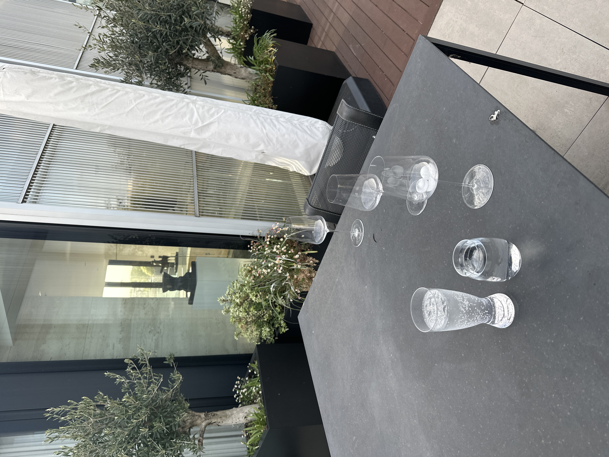 This media file captures an outdoor terrace or patio, likely in London, UK. In the foreground, a dark grey, rectangular table is laden with various clear glasses. These include several empty wine glasses, one containing ice cubes, and two tumblers filled with a clear, sparkling beverage. Adjacent to the table is a black outdoor speaker or chair with a perforated surface.
The setting features an assortment of potted plants in dark planters, including small olive trees and flowering bushes with delicate white and pink blossoms. A long, white, rolled-up fabric awning or canopy stretches along the perimeter, bordering the plants. In the background, a modern building with large glass windows and horizontal louvers is visible, with reflections of an interior space. The ground is composed of both wooden decking and light grey patio tiles.
The bright, natural lighting suggests it is daytime, under clear weather conditions. The scene appears peaceful and orderly, set up for relaxation or a quiet gathering. There is no discernible activity taking place, but the arrangement of glasses indicates recent or impending use. No visible text is present in the image.