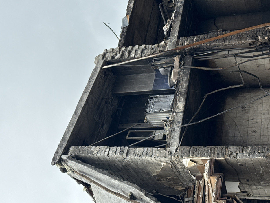 The photo depicts the ruins of a building, likely caused by destruction or fire. The exposed interior reveals a kitchen with blue tiles and damaged appliances. Debris and broken wires are visible, suggesting a collapse or significant structural damage. The perspective looks upwards, emphasizing the height and devastation. The sky is overcast.