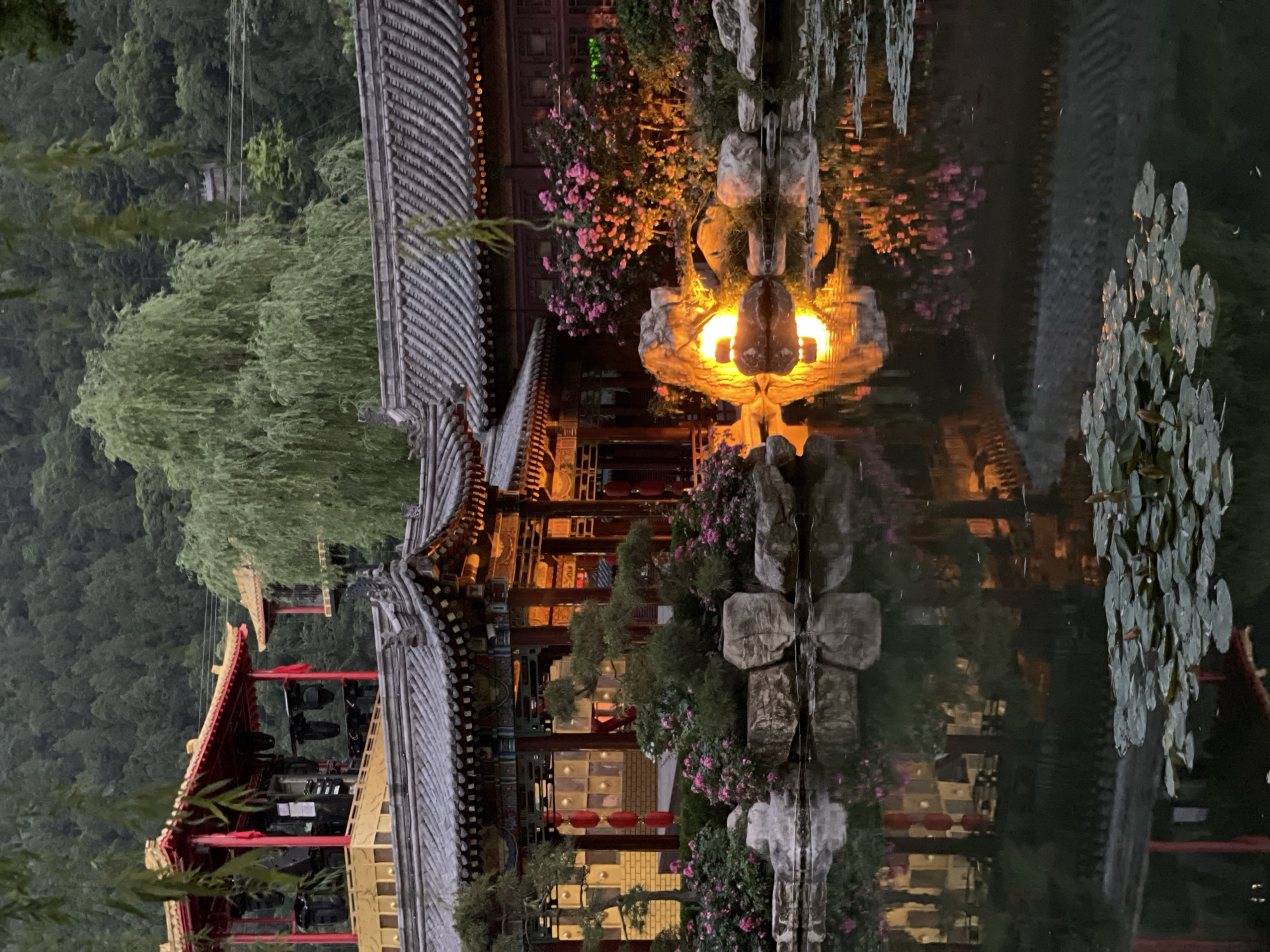 The image shows a nighttime scene in Lintong District, China, with a reflection of illuminated traditional Chinese architecture in a body of water. The structures feature gray tiled roofs, red and gold accents, and ornate details. A tree with drooping branches is situated on the left, and a rocky structure with a lighted element is visible. Pink flowers are also present. The reflection in the water creates a symmetrical effect, blurring the distinction between the real and reflected elements. The background consists of a densely forested hillside.