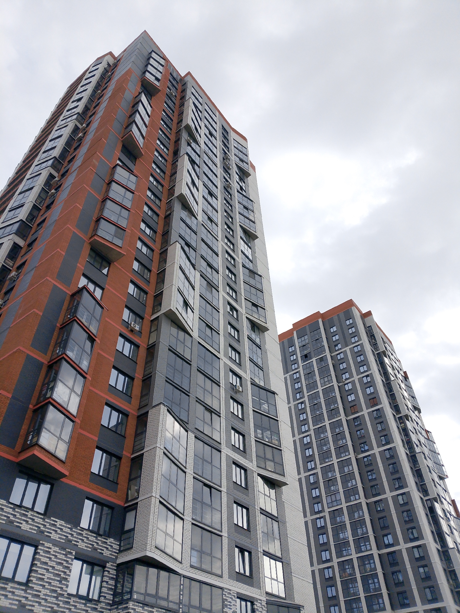 The image captures a low-angle view of two high-rise buildings against a cloudy sky. The building on the left features a combination of red brick and gray panels with protruding glass-enclosed balconies. The building on the right is predominantly gray with a similar architectural style. The buildings appear to be apartment complexes. The image was taken in Izhevsk, Russia.