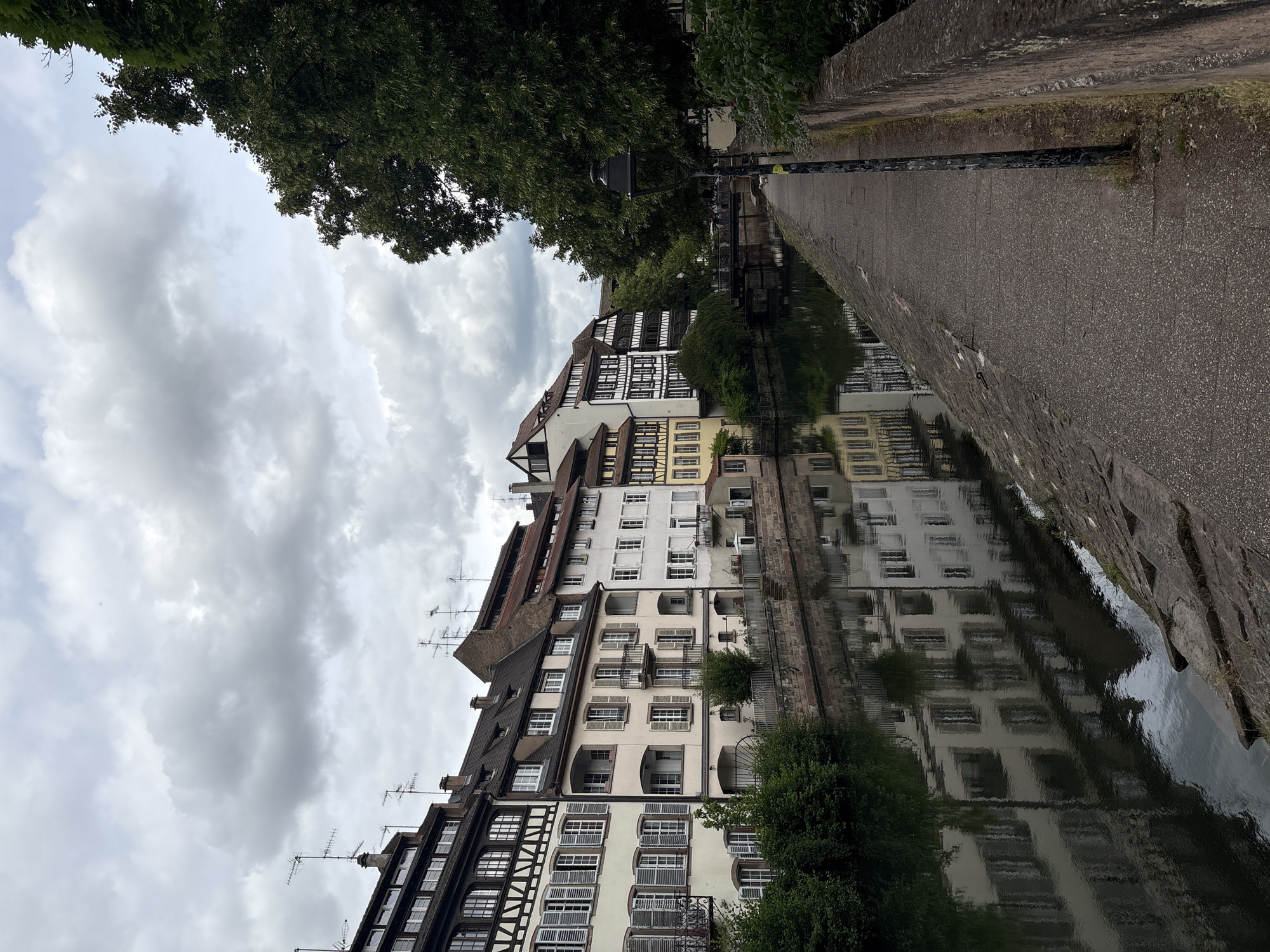 The image captures a canal lined with traditional Alsatian buildings in Strasbourg, France. The buildings exhibit characteristic half-timbered facades and colorful exteriors, with numerous windows and shutters. Several antennas are visible atop the roofs. The canal reflects the buildings and the overcast sky, which is filled with dramatic, grey clouds. On the right side of the canal, a cobblestone walkway runs alongside, bordered by a stone wall and lush green trees. A black lamppost stands on the walkway, and in the distance, a bridge spans the canal. A small white dog is visible on the walkway near the lamppost. The overall scene conveys a serene, historical urban landscape.