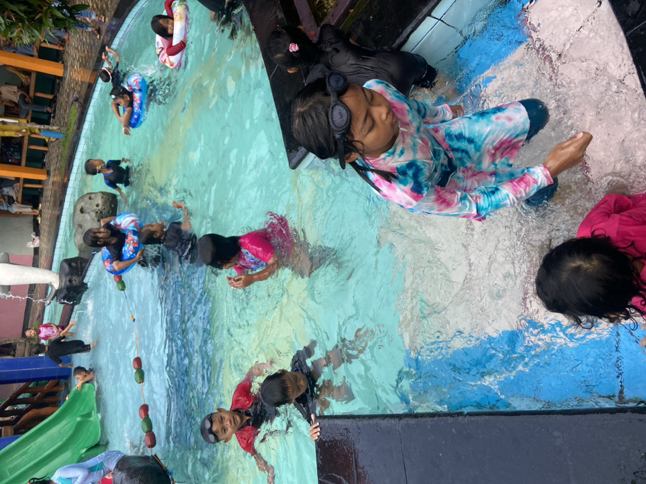 The image shows a swimming pool with multiple children swimming and playing in the water. The pool is located in the city of Kecamatan Tanggulangin, Indonesia, with coordinates of latitude -7.497 and longitude 112.684. The pool is surrounded by a concrete deck and a small area with grass and plants. There is a slide and a small waterfall entering the pool. There is also a rope with floating balls in the pool, and some children are using inflatable tubes. The children are wearing various swimming attire, including swimsuits, rash guards, and floaties. Some of them are interacting with each other while playing in the water. There is a girl in a pink and blue patterned swimsuit standing on the edge of the pool with her hand outstretched towards the water. Behind her, a group of children are enjoying the swimming pool. Some adults are sitting on the edge of the pool watching the children, but they are not visible in the picture. The pool is surrounded by a concrete wall and a fence. There is a small, light-colored building and a small garden nearby. The pool itself has a blue, tiled surface and is filled with clear water.