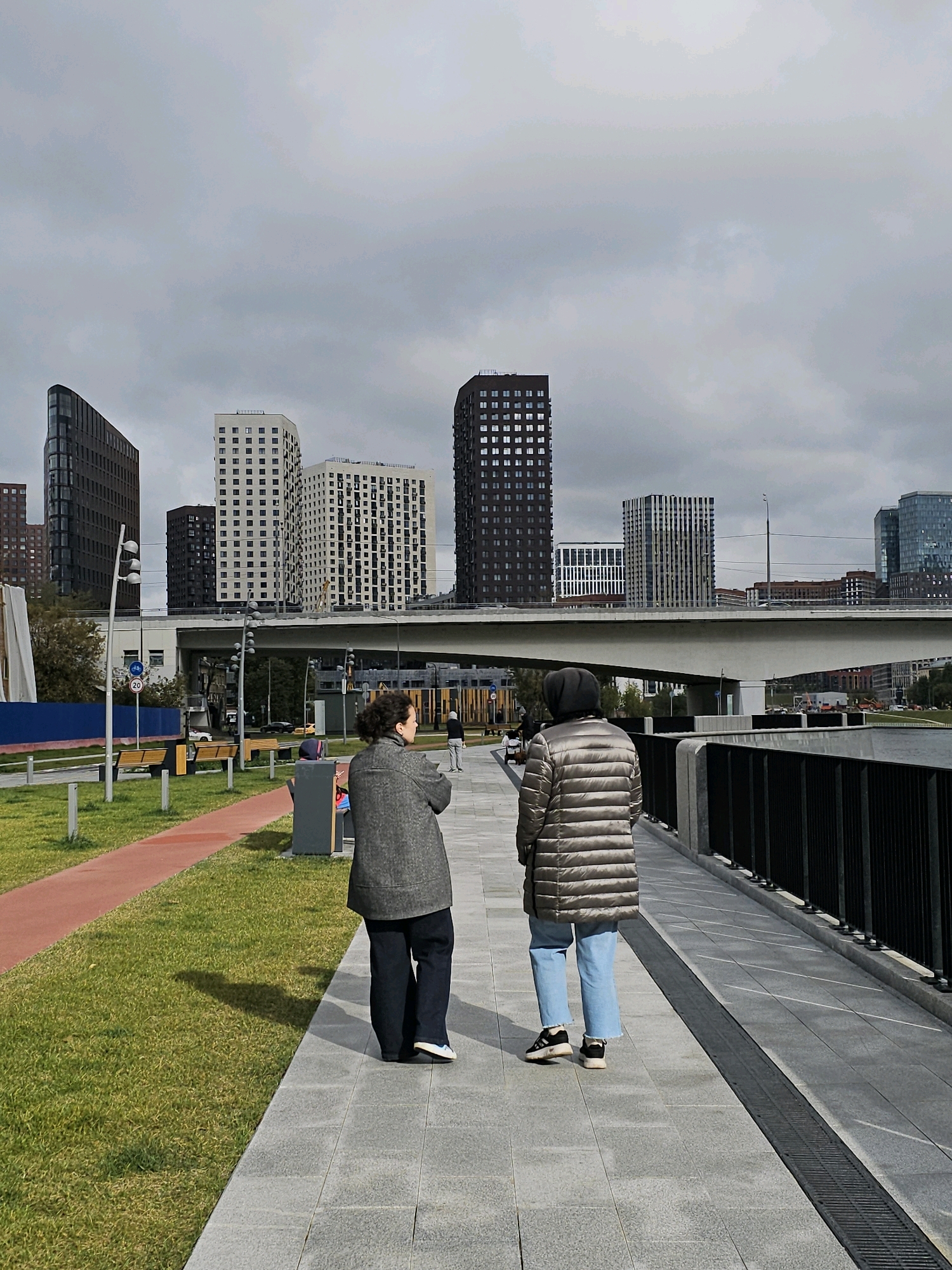 Two women walk along a paved waterfront promenade under a cloudy sky. The woman on the left wears a dark gray textured coat and dark trousers. The woman on the right is dressed in a puffed, light brown jacket and light blue jeans. Behind them, the promenade is bordered by a black metal fence and a wide body of water. In the background, a modern pedestrian bridge spans the water, with several contemporary high-rise buildings visible beyond it. The buildings are a mix of dark and light-colored facades with numerous windows. In the foreground, to the left of the women, is a grassy area with a red running track. Benches are visible in the grassy area. The scene appears to be in the Nagatino i-Land area of Moscow, Russia, a newly developed district known for its modern architecture and waterfront amenities.