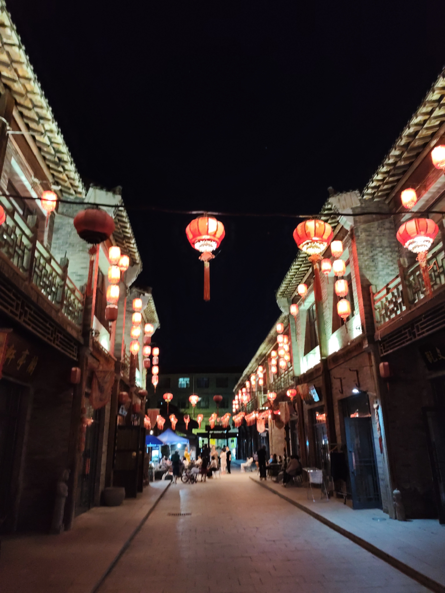 This image depicts a street scene at night in Lindian, China, likely a commercial or cultural area adorned with traditional Chinese red lanterns. The lanterns are strung across the street, illuminating the traditional-style buildings on either side. The architecture features multi-story structures with wooden balconies and tiled roofs, characteristic of older Chinese towns. Some buildings have ornate decorative elements.
The street itself is paved with stone or brick, and it appears to be a pedestrian walkway. Along the sides of the street, there are shops or restaurants with open entrances and displays. In the background, more lanterns are visible, as well as the shapes of other buildings and possibly some trees.
Several people are present, though their faces are not clearly discernible due to the low light and distance. Some individuals are seen walking, while others appear to be seated at tables, perhaps at outdoor dining areas. There are also some bicycles or carts visible. The overall atmosphere is festive and inviting, with the warm glow of the lanterns creating a unique ambiance. The sky above is dark, indicating it is well after sunset.