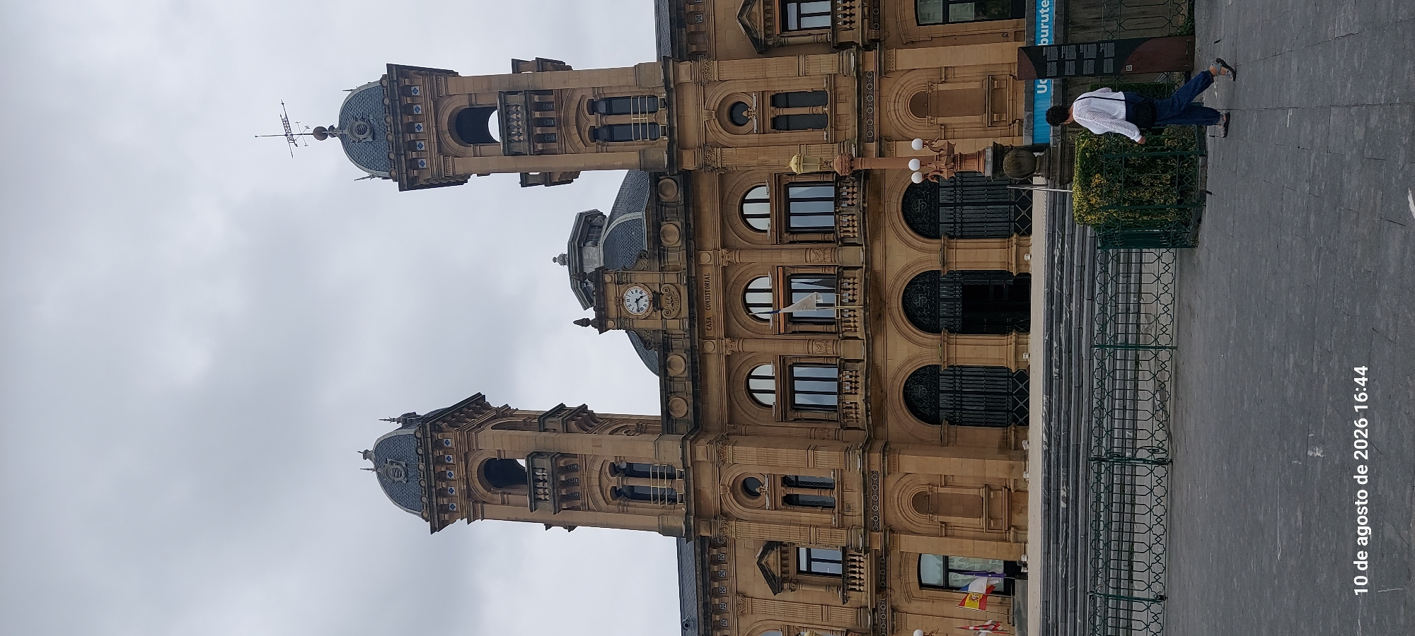 This image shows the Palacio de la Kursaal in San Sebastián, Spain, under a cloudy sky. The building is a grand, ornate structure made of tan stone, with two prominent towers topped with dark domes. A large clock is visible on the facade between the towers. In front of the building, there is a wide stone staircase leading up to arched doorways with decorative metal gates. A metal railing with a geometric pattern runs along the staircase. To the right of the staircase, a woman wearing a white sweater and blue jeans is walking away from the camera. She has a black bag slung across her shoulder. A lamppost with multiple globes stands to the right of the woman. In the foreground, the ground is paved with grey stone. Small flags are visible on the left side of the building. The date stamp at the bottom of the image reads "10 de agosto de 2026 16:44."