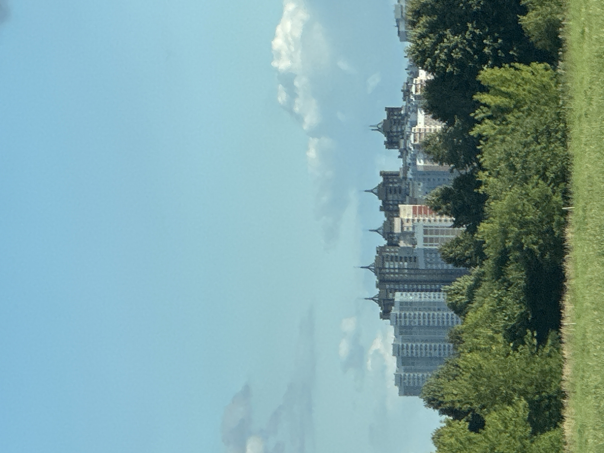 The image depicts a cityscape with a prominent blue sky dotted with white clouds. In the mid-ground, a cluster of apartment buildings of varying heights and architectural styles is visible. Some buildings are modern high-rises with repeating window patterns, while others have more distinctive decorative elements on their roofs. In the foreground, lush green trees form a dense canopy, with some lighter green foliage interspersed. Below the trees, a field of dry, light-colored grass stretches across the bottom of the frame. The overall scene suggests an urban environment bordered by green spaces.