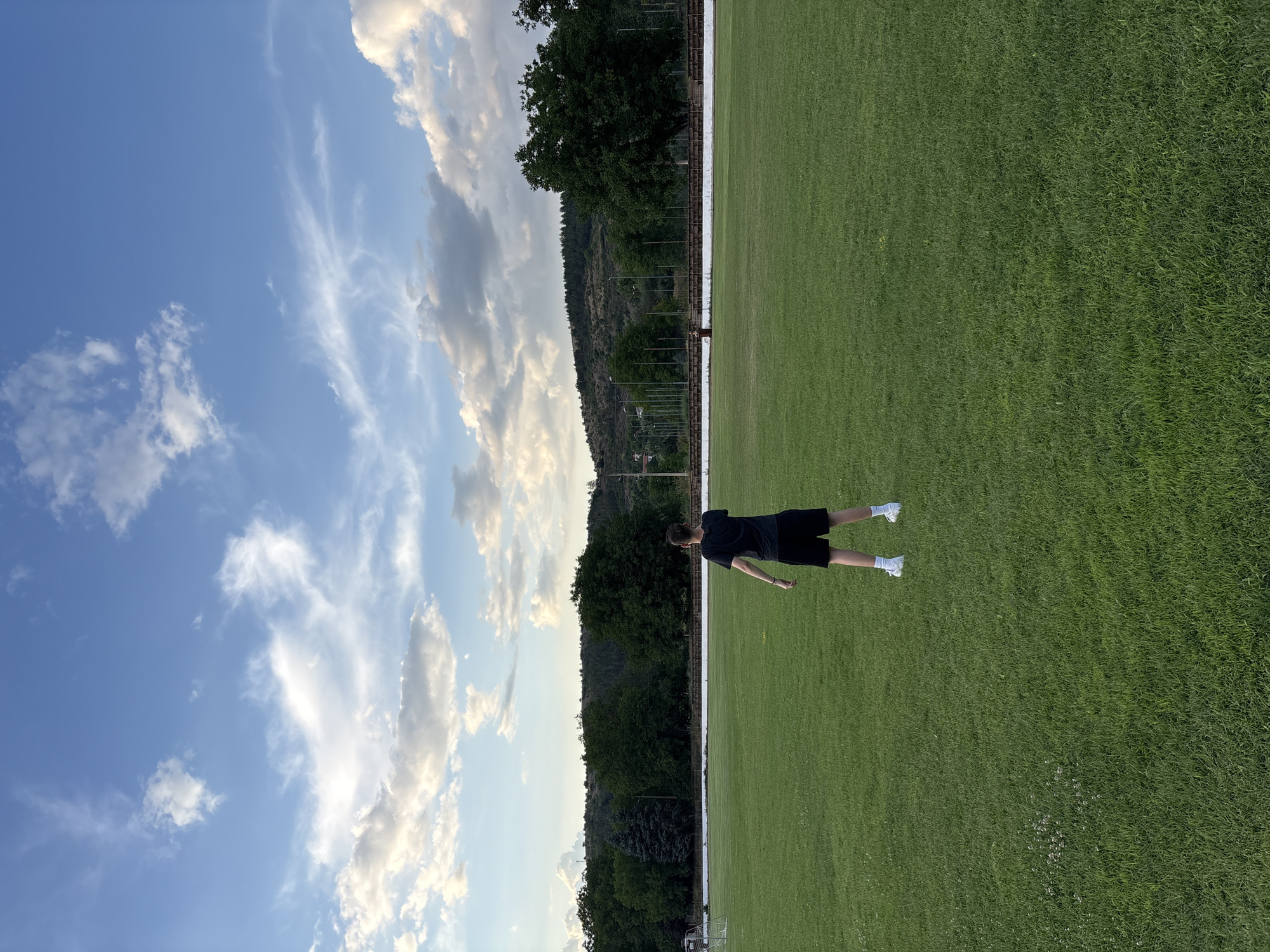 The image depicts a young person standing on a vibrant green grassy field, possibly a sports field, in Boboshevo, Bulgaria. The person is facing away from the camera, wearing a black t-shirt and black shorts, with white socks and shoes. The field is expansive and well-maintained, with a slight slope in the background. In the distance, a line of trees forms a natural backdrop, beyond which is a gentle hillside covered in vegetation. The sky above is a clear blue, punctuated by scattered white clouds, indicating fair weather. The overall setting appears to be an outdoor recreational area within the city.