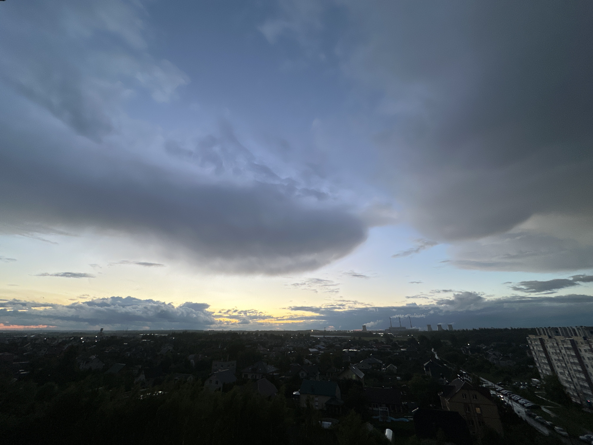 The image depicts an aerial view of a suburban landscape at dusk. A large portion of the frame is occupied by a dramatic sky filled with dark, heavy clouds that are beginning to break, allowing the warm hues of the sunset to filter through. Below, a dense residential area is visible, characterized by numerous houses with dark roofs and scattered trees. In the background, industrial structures with tall chimneys are prominent, with plumes of smoke or steam rising from them, suggesting a power plant. A multi-story apartment building is also visible on the right side of the frame. The lighting suggests either the approach or departure of a storm. The location is identified as Shchomyslitski Sel'ski Savet, Belarus.