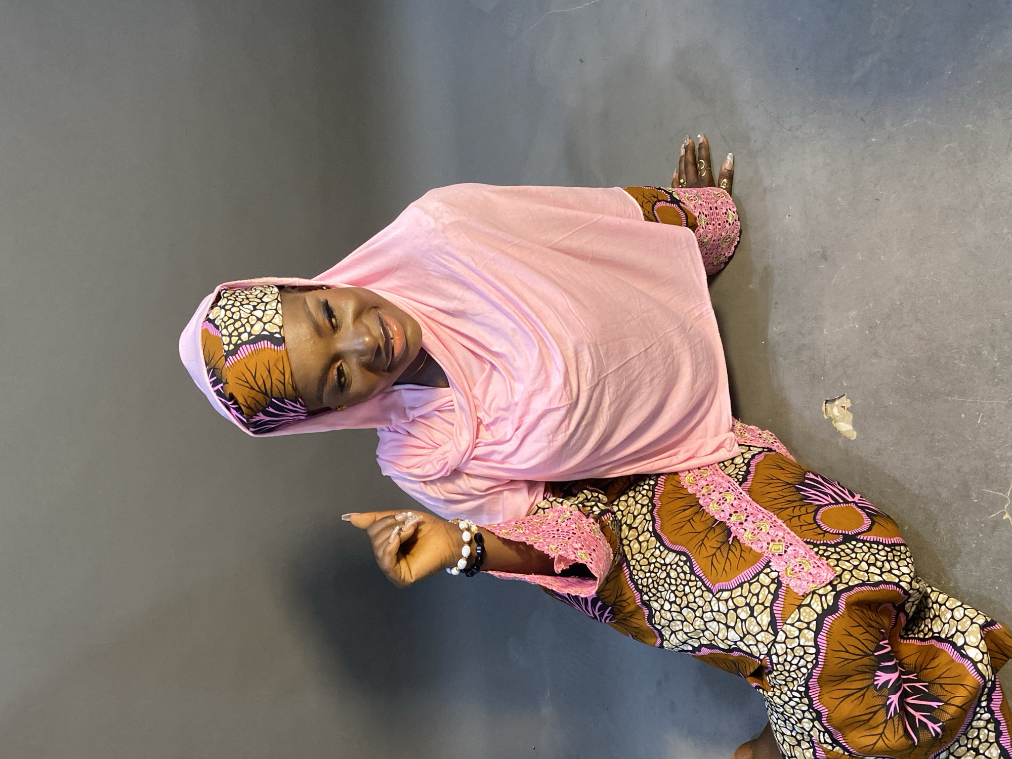 A vibrant medium shot captures an African woman posing indoors against a plain dark grey background. The image is rotated 90 degrees counter-clockwise. The woman, who has dark skin, is smiling broadly at the camera. She wears a light pink hijab that covers her head and drapes over her shoulders, and a patterned dress featuring brown, pink, black, and white motifs, likely an Ankara fabric. The sleeve of her dress has an intricate pink lace cuff.
Her right hand is raised and pointing, adorned with white and black beaded bracelets on her wrist and rings on her fingers. Her nails are long, manicured, and painted. Her left hand is also visible, with similarly manicured and painted nails, and rings on her fingers and toes. The woman's expression conveys happiness and warmth. The setting appears to be a simple indoor space with a concrete or tiled floor. No text is visible in the image. The attire is consistent with fashion often seen in West Africa.