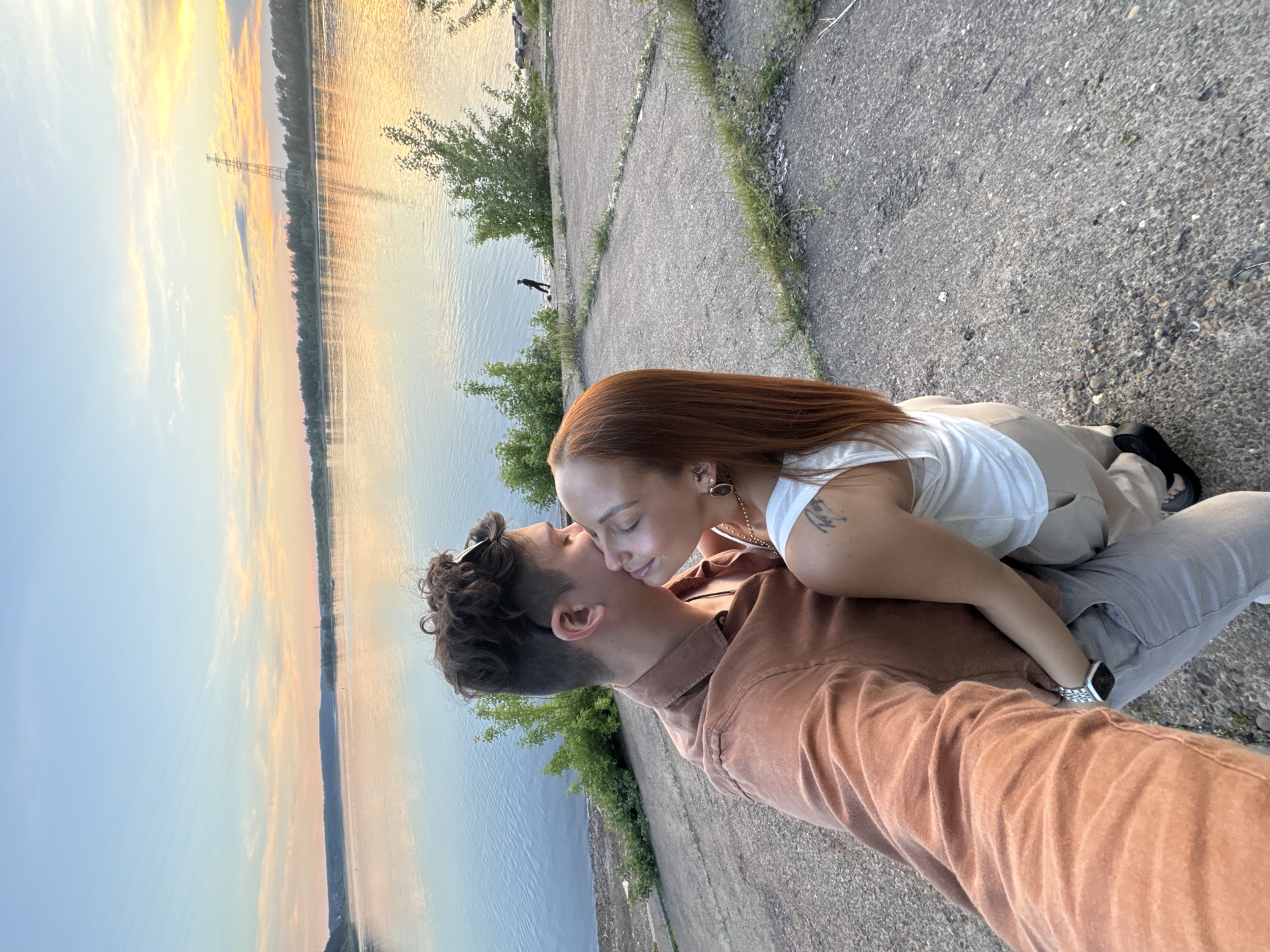 A couple embraces by a body of water at sunset, with a serene sky painted in hues of orange, pink, and blue. The woman, with long auburn hair, leans her head against the man's cheek, both with eyes closed in a moment of affection. The man, wearing a brown button-down shirt, has his arm around her waist. They are standing on a textured concrete surface, possibly a pathway or embankment. In the distance, a lone figure walks near the water's edge, and a tall transmission tower stands against the vibrant sky. The location is identified as Noviy, Russia, suggesting a riverside or lakeside setting within the city.