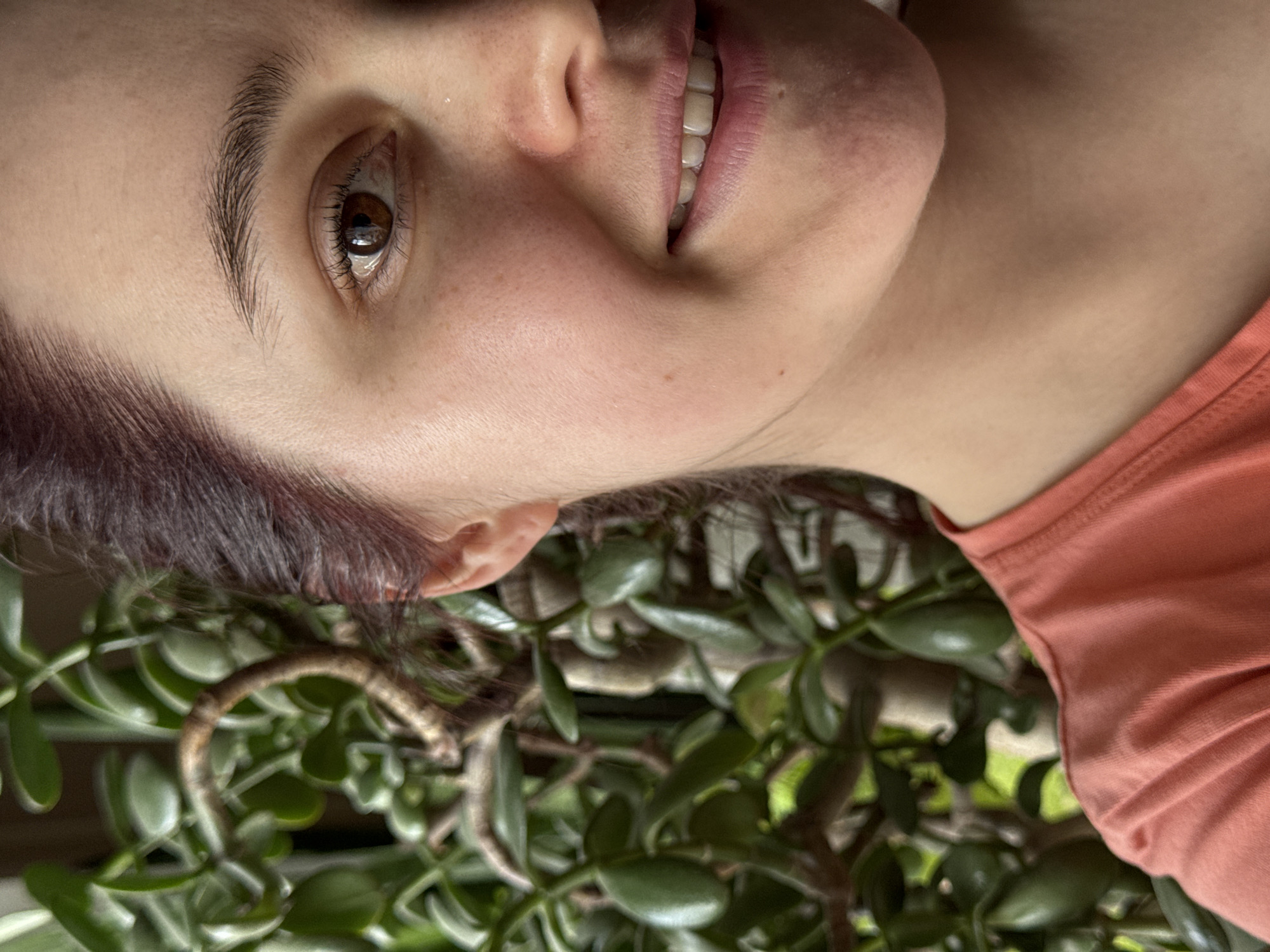 The media file is a close-up portrait of a person, rotated 90 degrees clockwise. The subject is a young individual with fair skin, dark brown eyes, and dark hair that has a subtle reddish tint. They are smiling broadly, revealing their upper teeth, and looking directly towards the viewer, conveying an emotion of happiness or amusement. Visible details include well-defined eyebrows and eyelashes.
The person is wearing an orange or coral-colored tank top or t-shirt, visible on the right side of the frame (when viewed in correct orientation). The background features a lush, green plant, identifiable as a jade plant (Crassula ovata), with its distinctive thick, fleshy, oval leaves and sturdy branches. The plant is positioned behind and slightly below the person's head, filling the lower and left sections of the image. The lighting appears natural, possibly from a window or an indoor setting. There is no visible text. The scene takes place in Izhevsk, Russia.