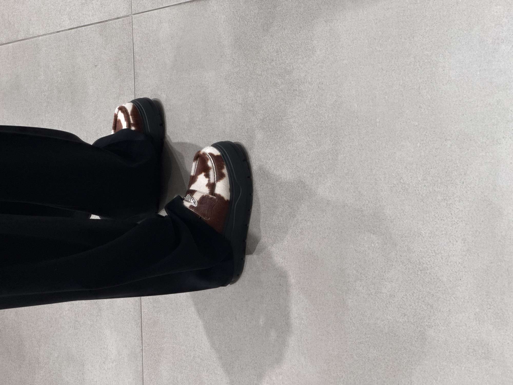 The image shows a top-down view of a person's legs and feet standing on a light gray tiled floor. The person is wearing black pants and brown and white cow-print loafers with a thick black sole. The environment appears to be indoors, possibly a public space with tiled flooring. There are no other discernible people or objects in the immediate vicinity. The image does not contain information to determine if the image was taken in Dubai, United Arab Emirates.