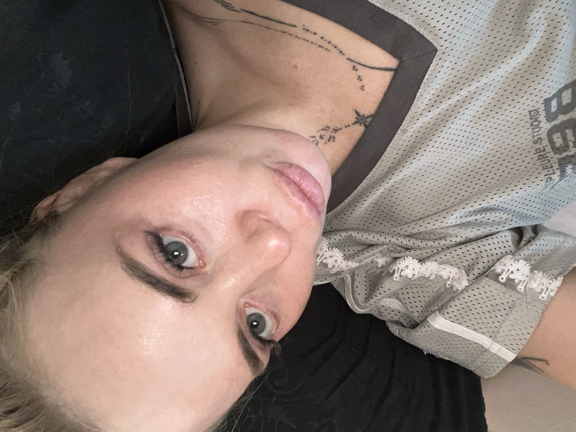 This is a close-up, vertically oriented image of a woman's face and upper torso. She is lying down, with her head tilted upwards and to the left. Her eyes are open and looking towards the camera. She has light skin, blonde hair, and is wearing makeup, including dark eyeliner and mascara, and what appears to be lip gloss.
She is wearing a light grey, mesh t-shirt with a dark grey neckline. The t-shirt has a lace trim along the sleeves and a white stripe detail. Visible on the fabric of her shirt is text, appearing to read "BEPURE STUDIO". The woman has tattoos on her neck and shoulder, with visible ink lines forming abstract shapes and a star-like design.
The background is dark and indistinct, suggesting an indoor setting with low lighting. The overall mood is candid and intimate, with the subject's direct gaze creating a sense of connection with the viewer. The lighting appears to be artificial, highlighting the texture of her skin and the details of her makeup and clothing.