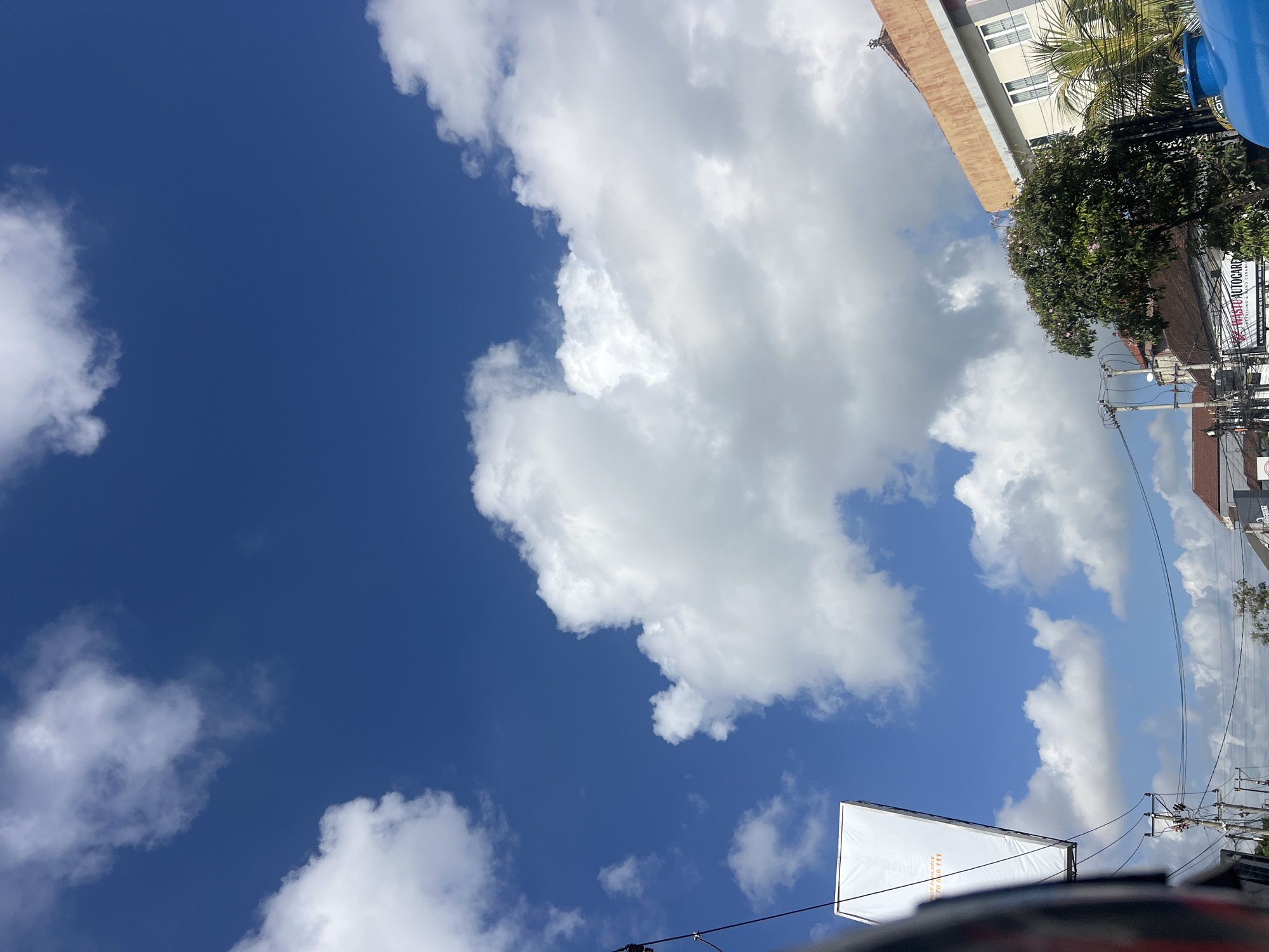 The image depicts a street scene in Denpasar, Indonesia, under a bright blue sky filled with fluffy white clouds. In the foreground, a portion of a dark object, possibly a vehicle, is visible on the left. A large, rectangular white billboard advertising "SPACE AVAILABLE" and a phone number is prominently displayed, suspended by wires. Electrical poles and wires crisscross the middle ground of the image, hinting at the urban setting. Several buildings with reddish-brown tiled roofs and light-colored walls are visible. One building on the right features white-framed windows and a prominent orange-brown roofline. A lush green tree with some pink blossoms stands in front of one of the buildings. In the lower right corner, a large, bright blue cylindrical object, likely a water tank, is partially visible. The overall scene suggests a typical street view in a populated area of Denpasar on a sunny day.