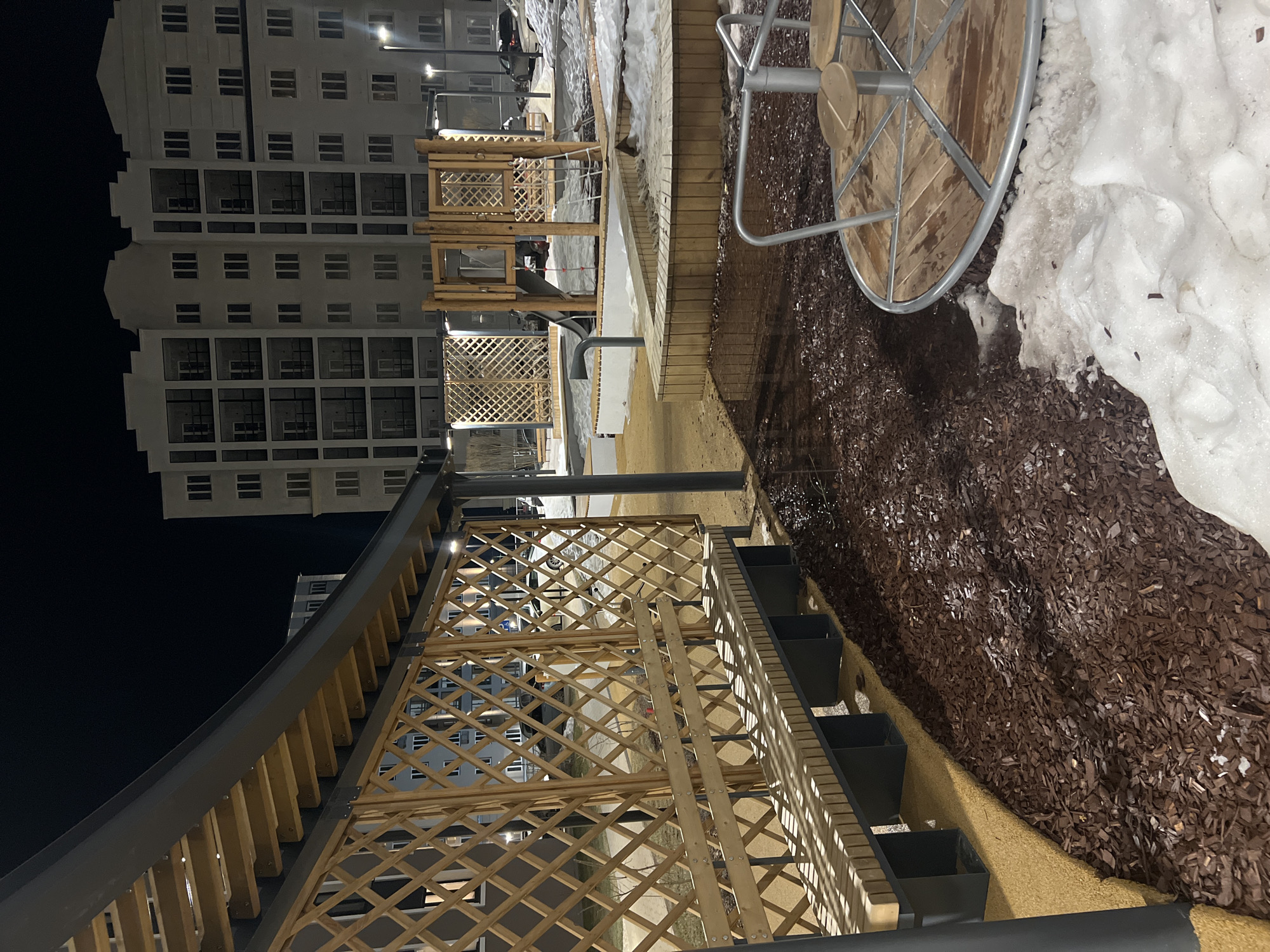 The scene depicts a modern outdoor playground or park area at night, located in Unknown, Unknown. No individuals are visible. In the immediate foreground, a circular wooden merry-go-round with a metallic frame is partially visible, surrounded by dark brown wood chip mulch and encroaching patches of white snow. To the left, a large wooden pergola features a dark, curved roof, lattice-patterned walls, and an integrated wooden bench. The ground directly beneath the pergola is covered in a light brown granular surface.
Further into the midground, a complex of wooden playground structures is present, including a slide, climbing elements, and raised platforms. These structures are primarily situated on dark brown wood chip mulch, with additional snow accumulation in various sections. The background features a large, light-colored multi-story residential building with numerous windows and balconies. Several streetlights illuminate the area, casting shadows and highlighting the textured surfaces of the playground equipment, mulch, and snow. Additional buildings, parked vehicles, and snow-covered ground are discernible in the far background. The sky is dark, indicating nighttime conditions.