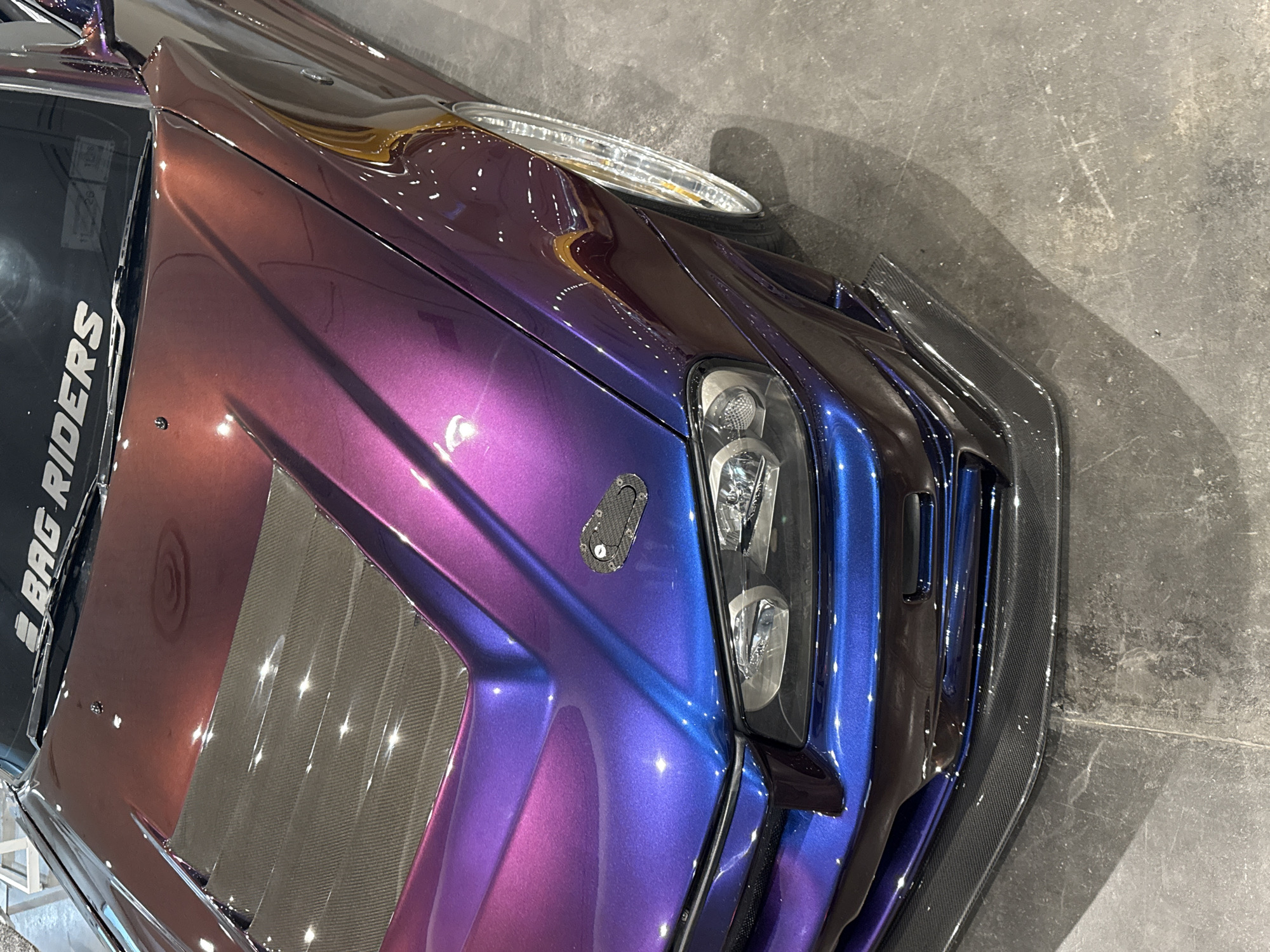 Here is a forensic description of the image:
The image shows a close-up, angled view of the front portion of a custom car. The car's paint job features a color-shifting effect, transitioning from a brown-purple hue at the top to blue in the lower right portion near the headlight. The headlight is multi-lensed. A black carbon fiber front splitter and a carbon fiber vent on the hood are visible. The windshield displays white lettering, part of which reads "BAG RIDERS". The car sits on a concrete-like surface, possibly the floor of a garage or exhibition space. There is no evidence in the image to connect the car to the city of New York. No people are visible in the image.