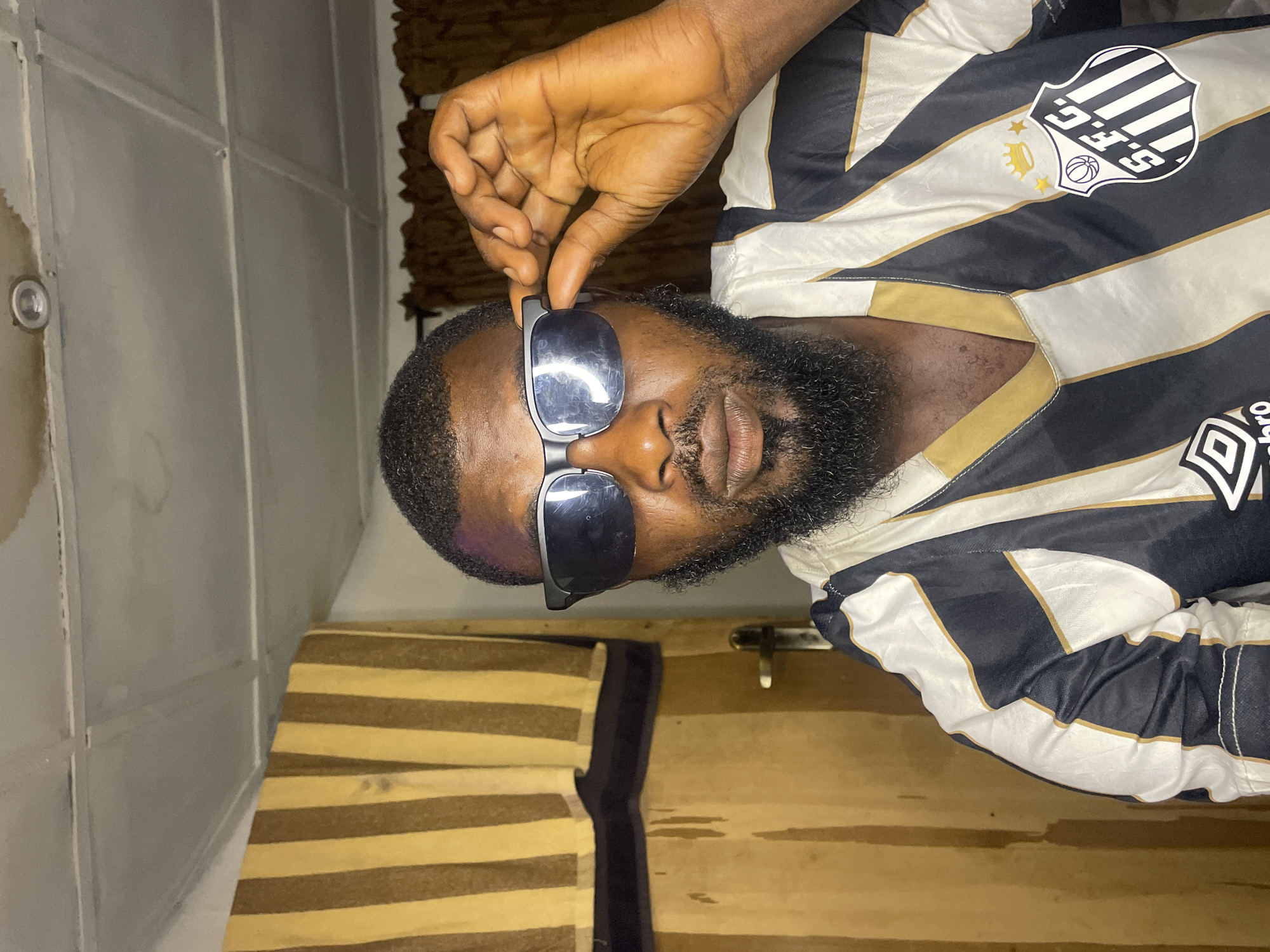 A bearded man with dark skin is holding his sunglasses up, revealing his eyes. He is wearing a black and white striped shirt with a logo on the upper left that includes the letters "S.F.G.". The background includes a white wall with visible seams, a wooden structure with a hasp, and a horizontally striped cushion. The location is assumed to be Jos, Nigeria based on request.